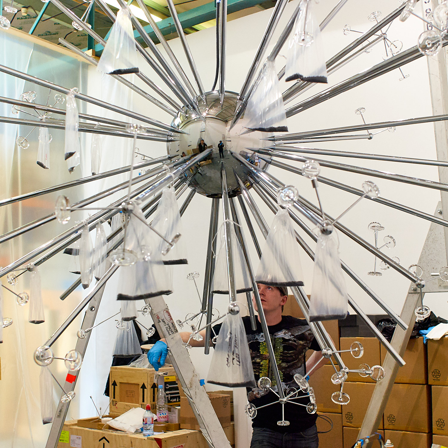 Person working on large, metallic spherical art installation with radiating rods in a workshop setting.