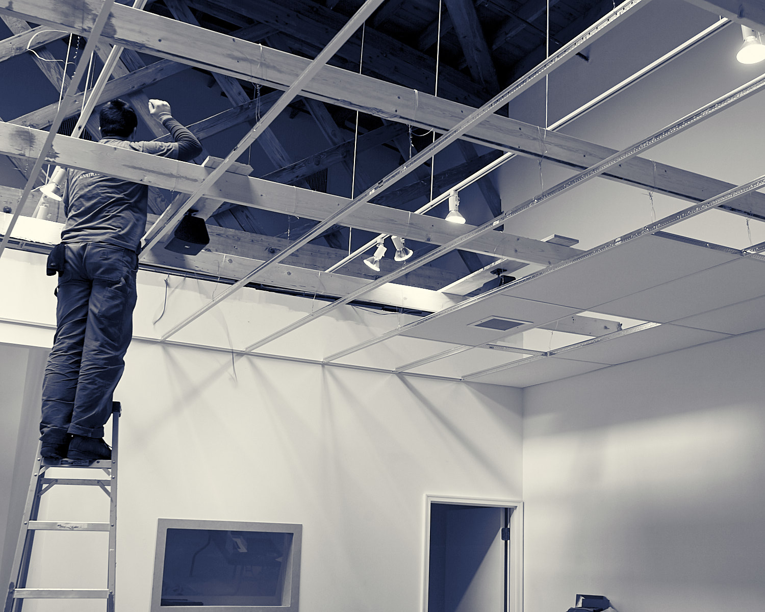 Person on ladder installing or repairing suspended ceiling with exposed beams in a room.