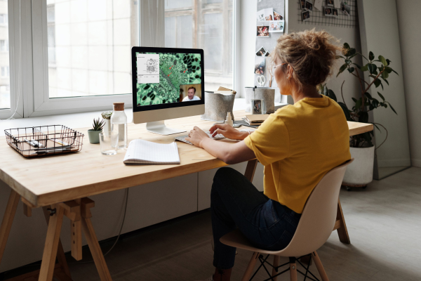 A woman sitting at a wooden desk, engaging in a video call on a desktop computer, in a bright room with natural light coming through a window.