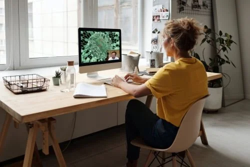 Girl in yellow T-shirt and hair up, sat at desk in clean modern room, learning online.