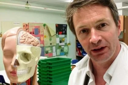 Link to Who Am I, photo of Tom in a biology lab, wearing a white coat and with an anatomy model head