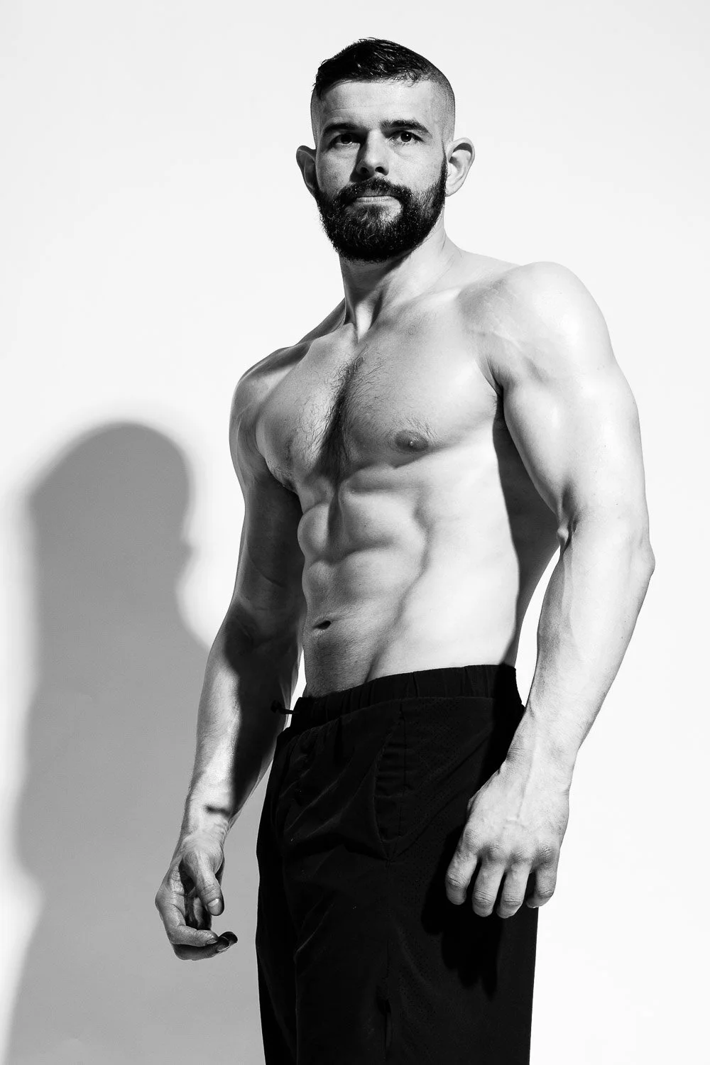 Black and white photo of a muscular, shirtless man with a beard and short hair, standing against a plain background.