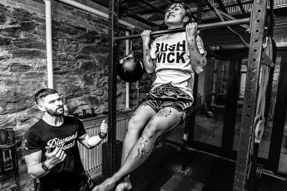 Two people exercising in a gym, one performing a pull-up while the other provides support; both appear focused and engaged in fitness.