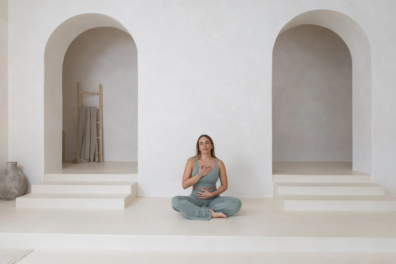 Mara sitting cross legged on the floor of a studio room with two arches, one hand resting on her heart