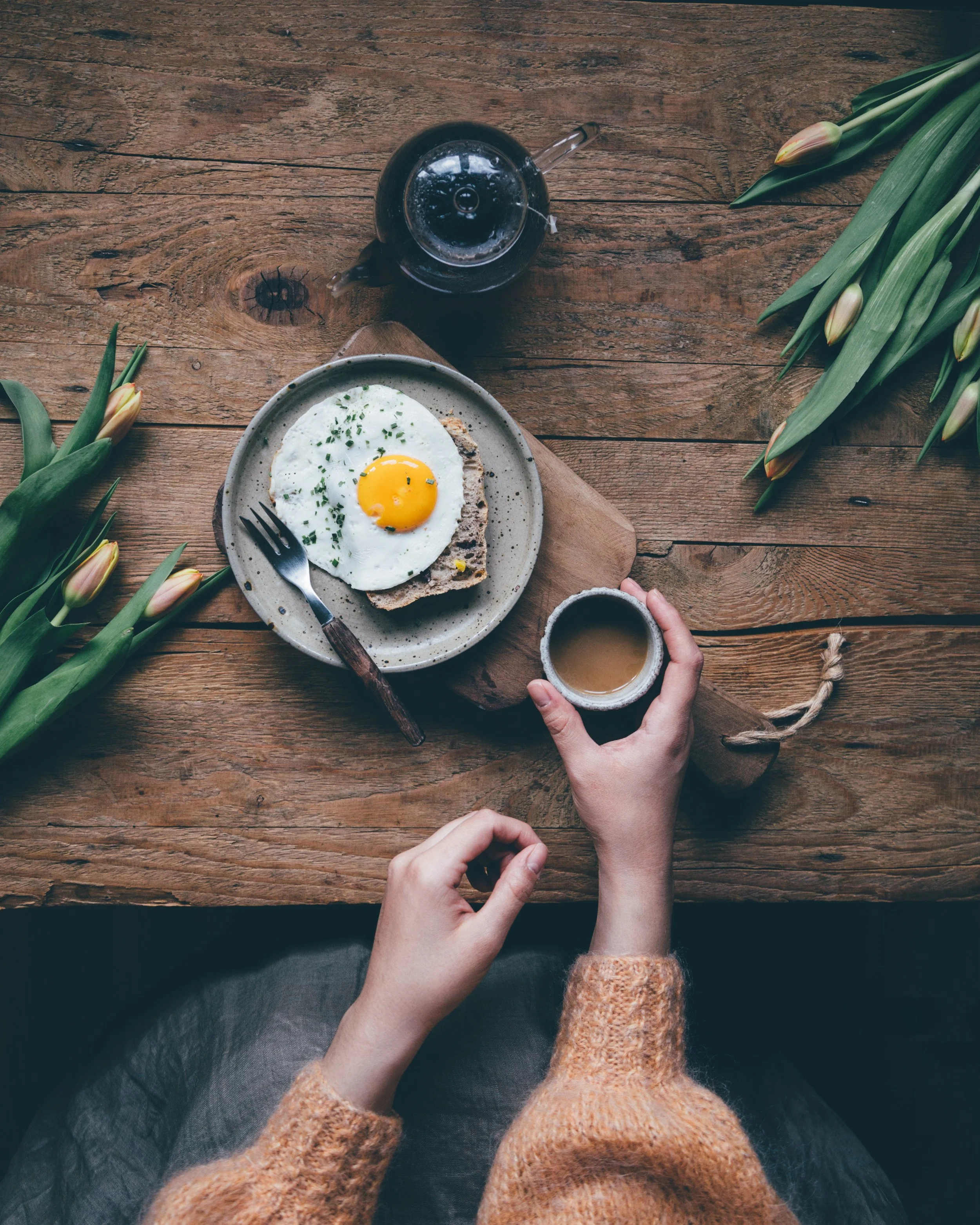 A wooden table is set for breakfast. There is a pot of coffee, tulips, and a piece of toast with a fried egg on top. A woman wearing a sweater is holding her coffee cup.