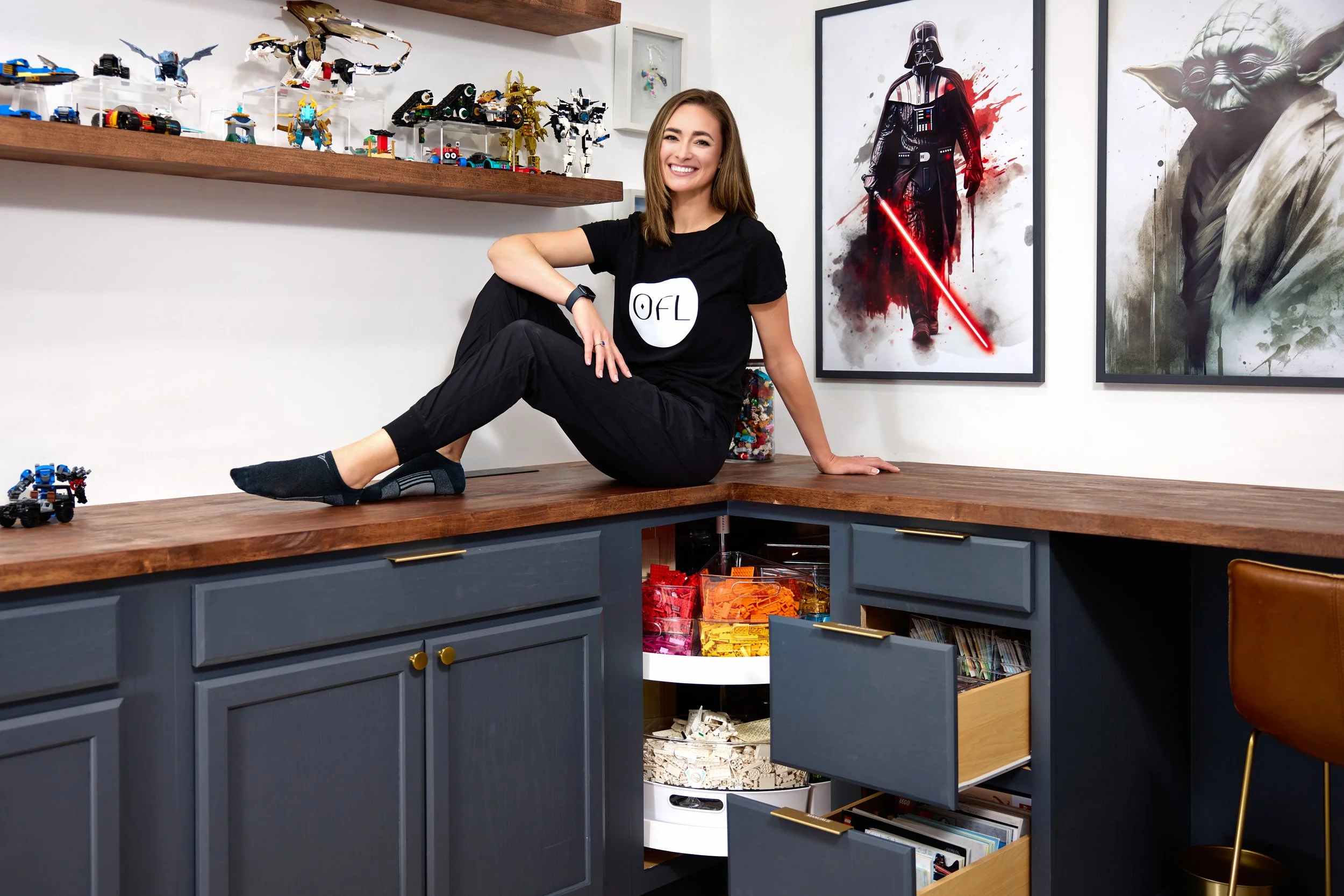 A woman with long brown hair smiling, sitting on a wooden countertop with blue cabinets below, in a room decorated with Star Wars and LEGO art. Shelves with LEGO sets are visible above her, and her t-shirt has the letters 'OFL' on it. An open cabinet shows organized LEGO bricks, and framed Star Wars posters are on the wall behind her.
