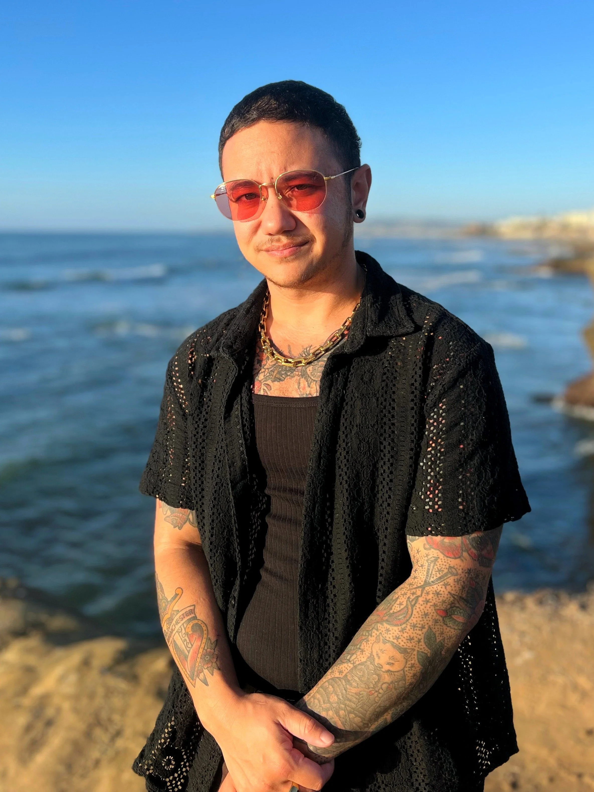 Portrait of someone with rose-tinted sunglasses wearing black clothes against a background of the ocean and cliffs at sunset.