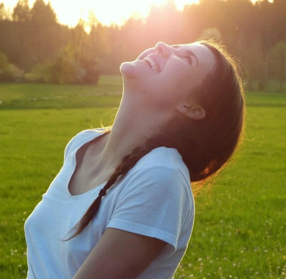 Une femme libre et vêtue d'un chandail blanc regarde vers le ciel avec un grand sourire