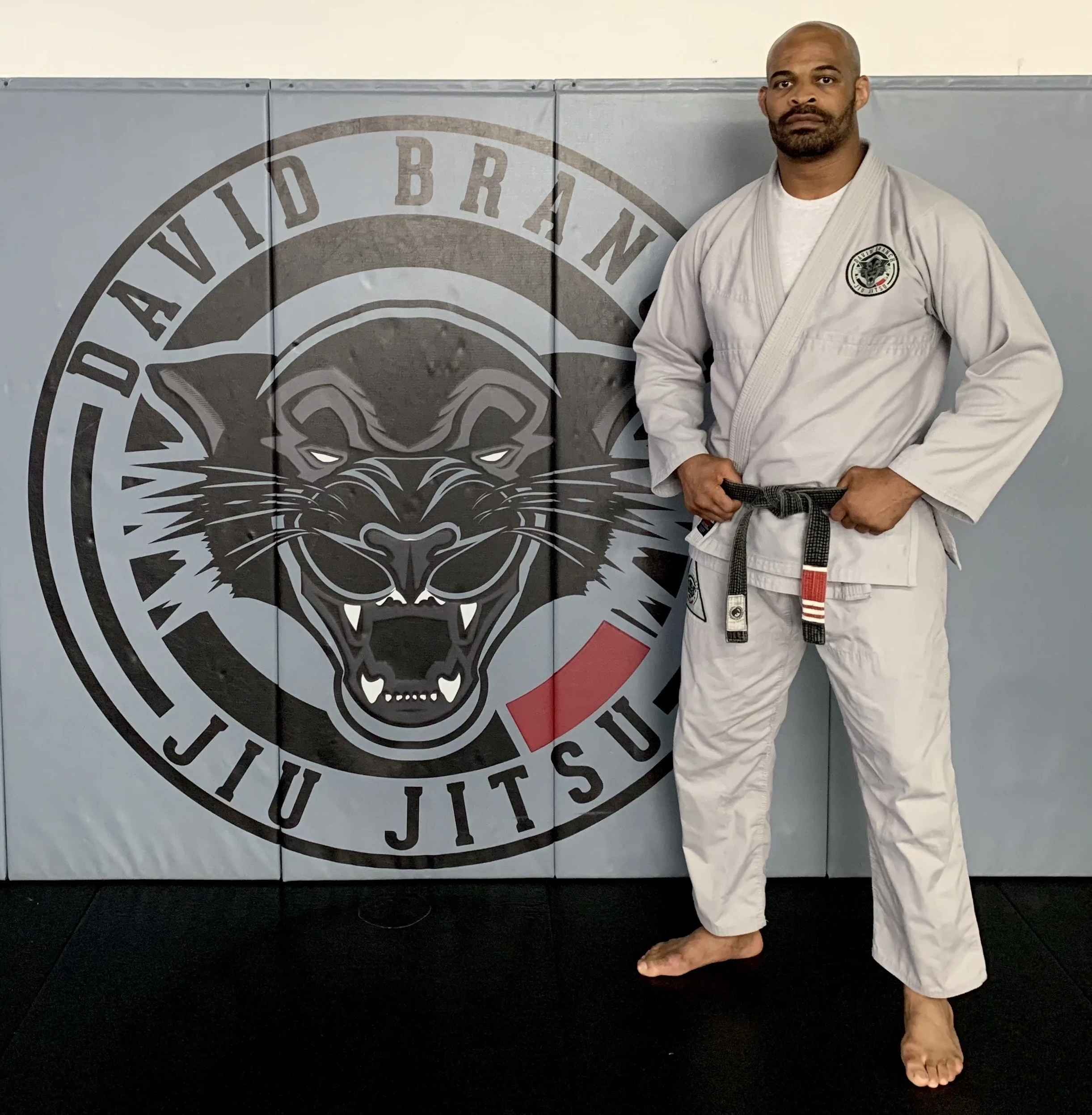 Gym owner, Professor David Branch, 2nd degree black belt, standing in front of the David Branch Jiu Jitsu logo in the gym