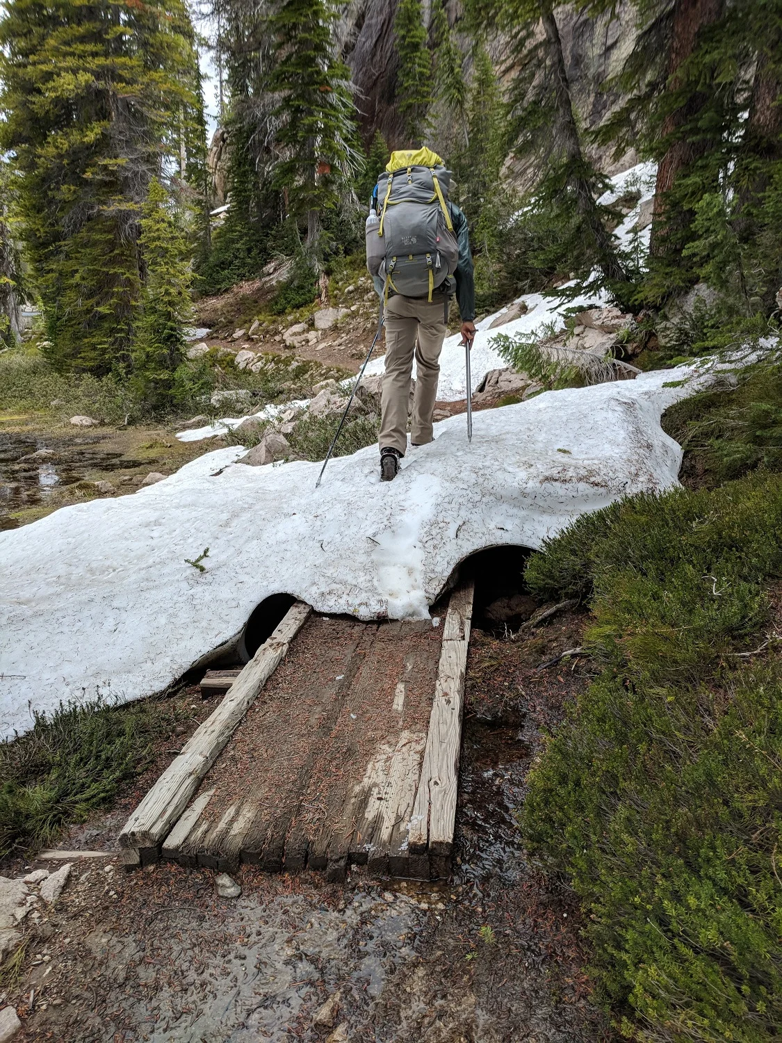 Andrew crossing a snow bridge over a wooden bridge over the soppy ground at the edges of Imogene Lake's marshy inlet