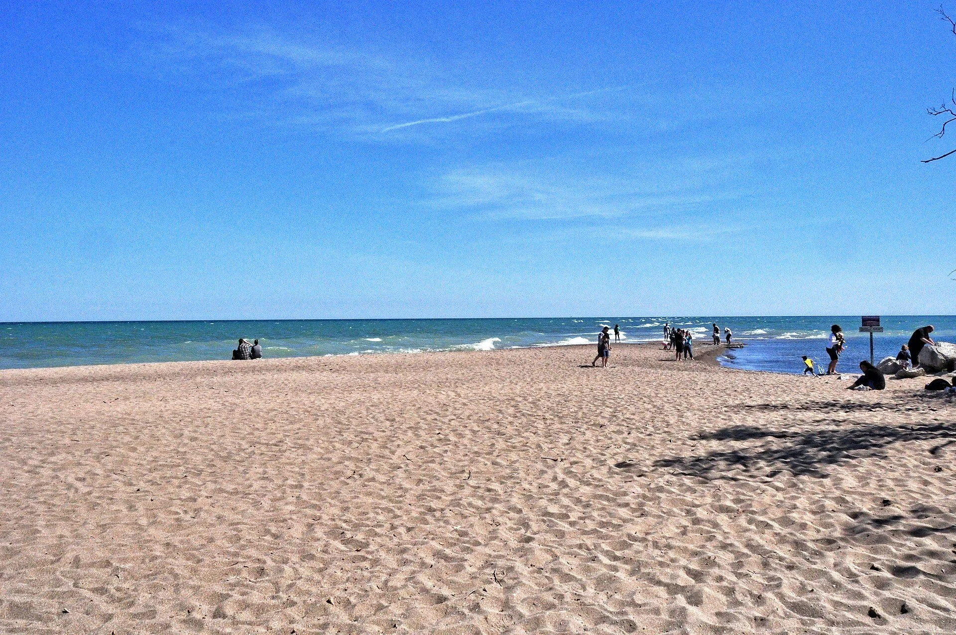 A wide sandy beach running out to Lake Erie at the Tip of Point Pelee under a bright blue sky, with people walking near the water.