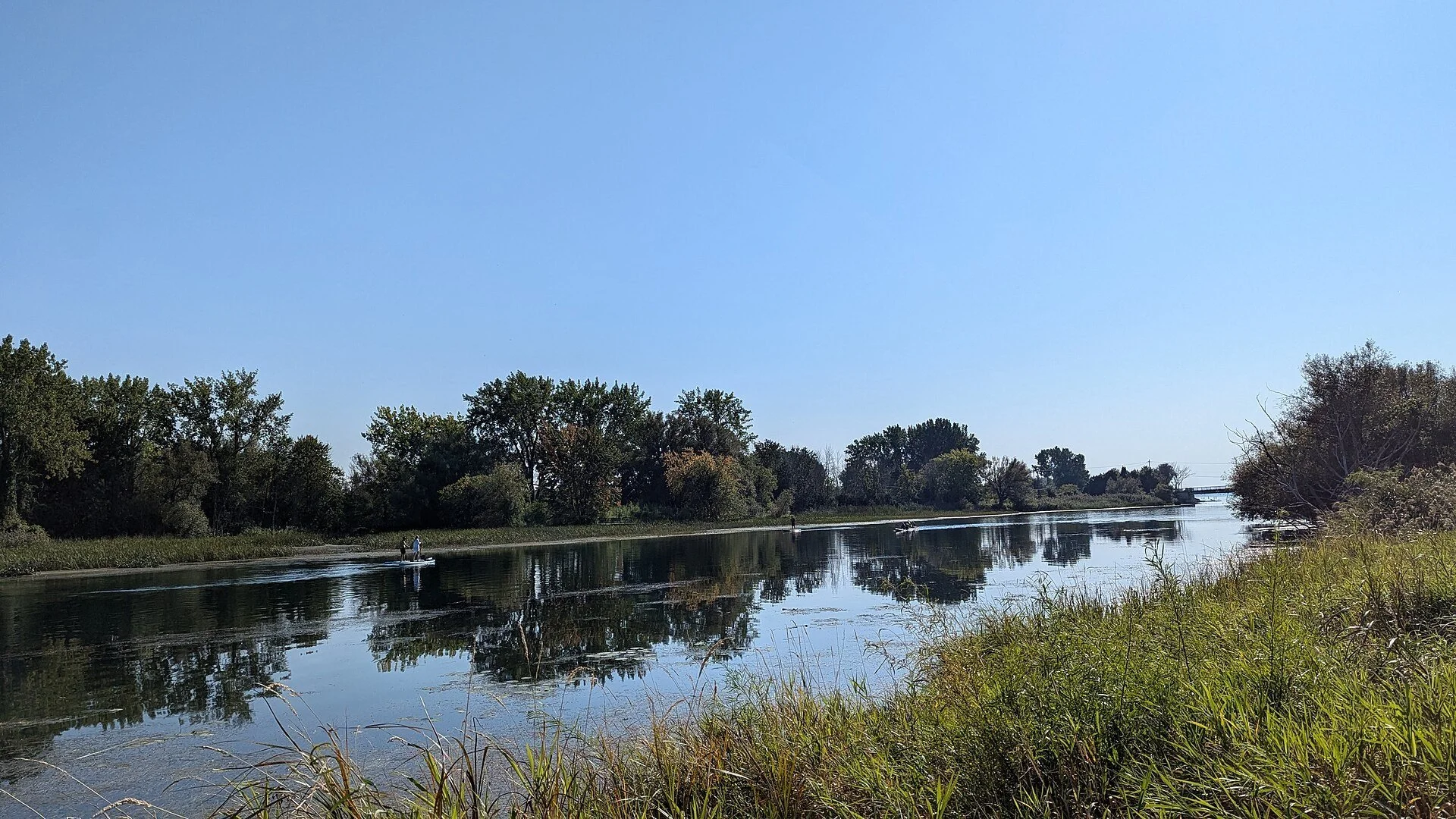The calm Châteauguay River reflecting a line of trees near Île Saint-Bernard under a clear blue sky.