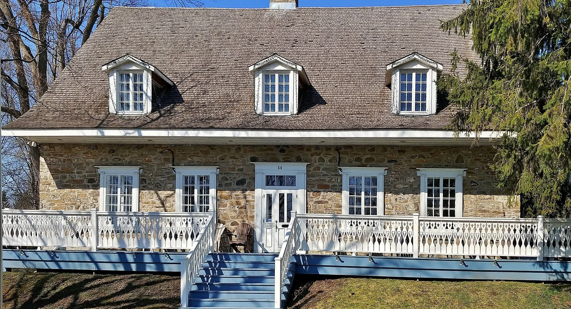 The front of Maison LePailleur, an eighteenth century stone house with dormer windows and a white veranda in Châteauguay.