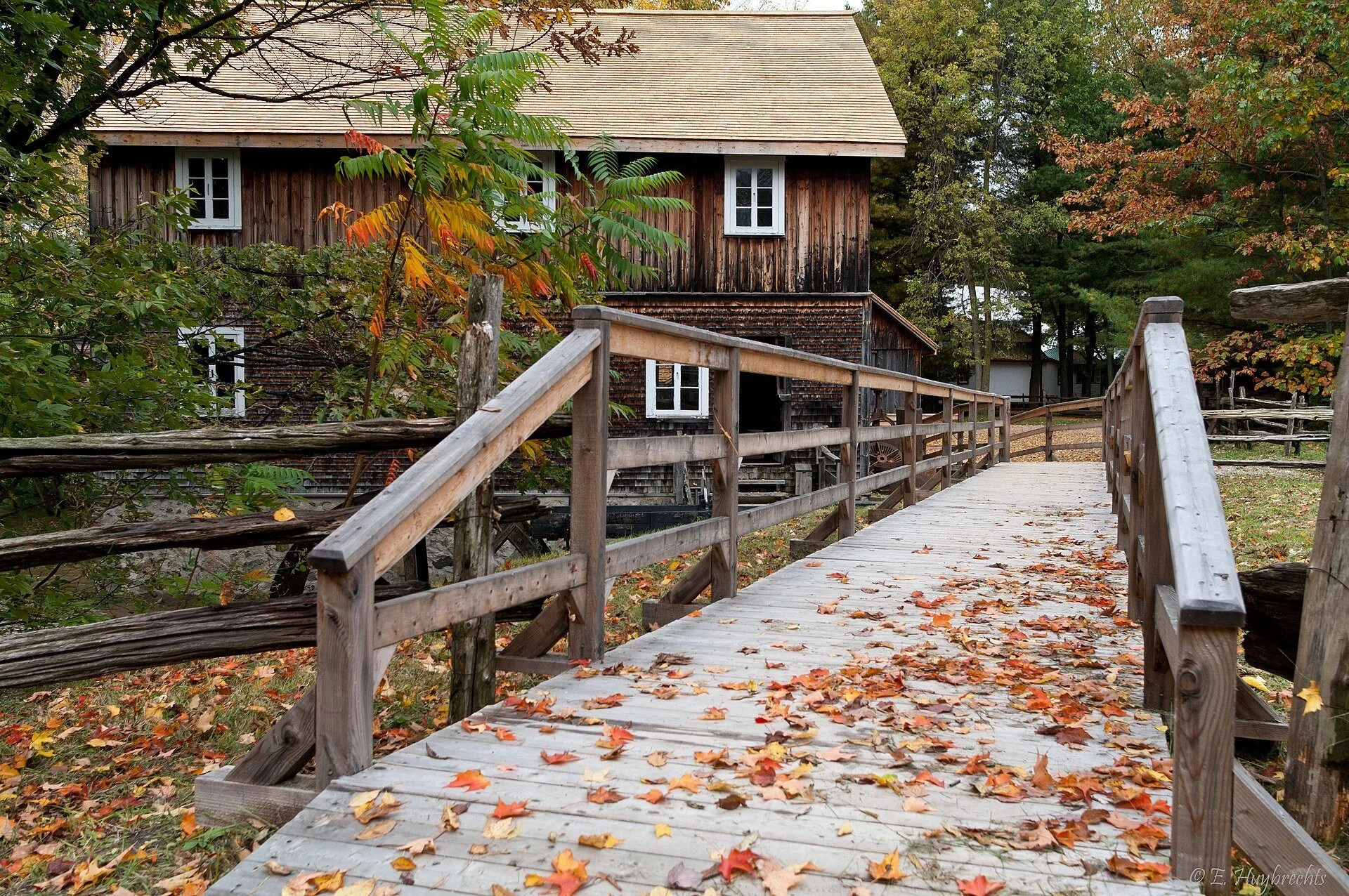 A wooden footbridge covered in fallen leaves leading to an old timber mill at the Village Québécois d'Antan in Drummondville.