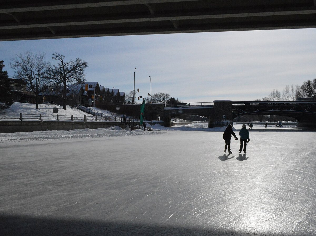 Two skaters glide side by side on the frozen Rideau Canal Skateway in Ottawa, seen from beneath a bridge on a sunny winter day.