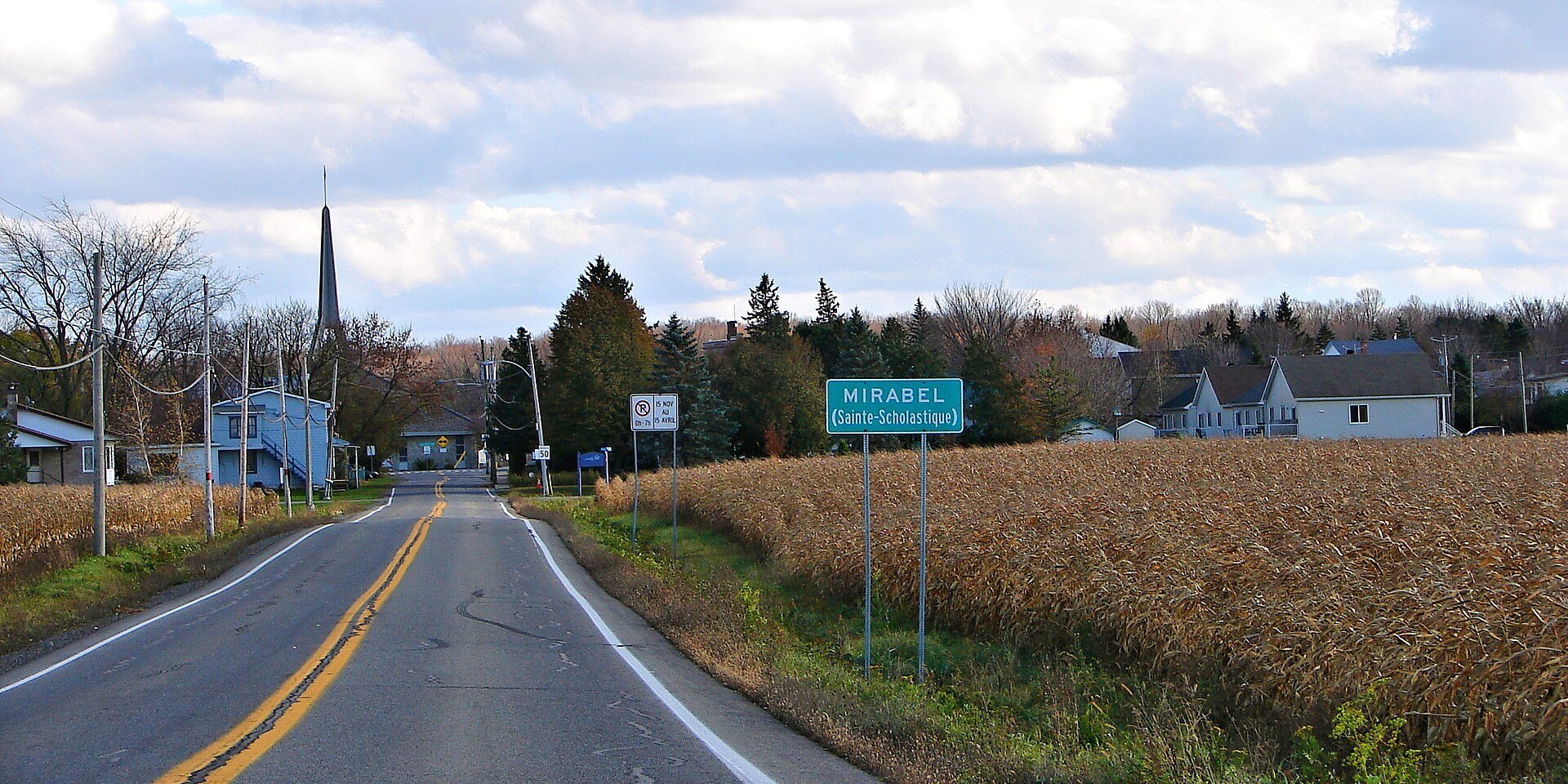 A country road between farm fields leads into the village of Sainte-Scholastique in Mirabel, Quebec, with the church spire ahead.