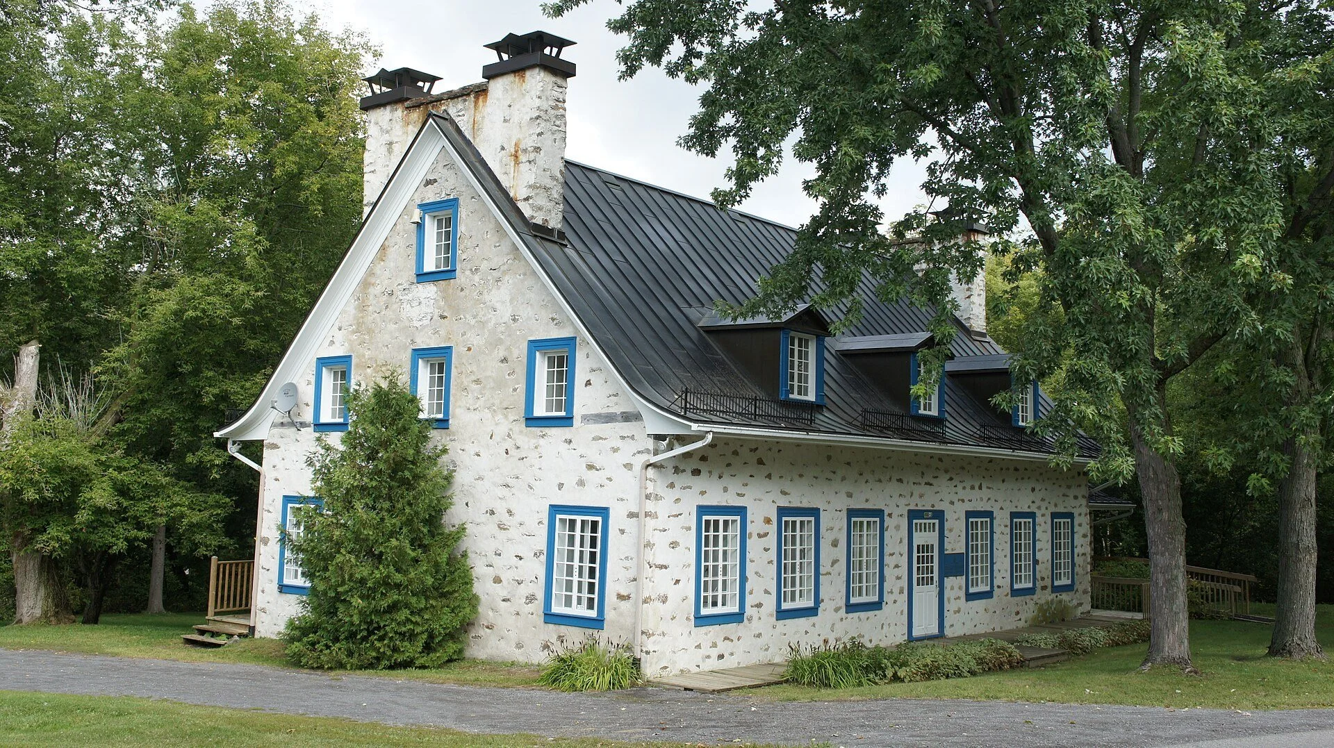 The stone Manoir de Belle-Rivière with blue shutters under tall trees on the Belle-Rivière estate in Mirabel, Quebec.