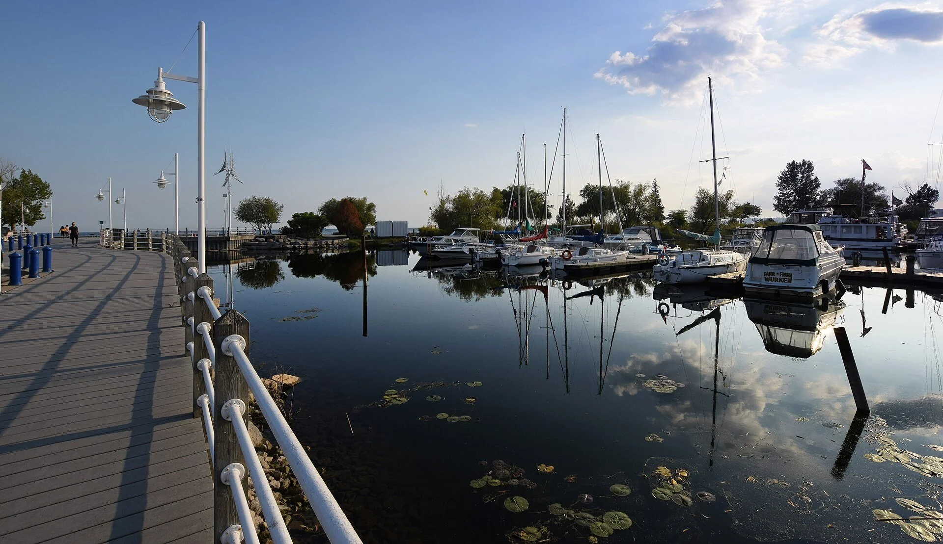 A boardwalk with lamp posts and railings beside boats reflected in Frenchman's Bay, Pickering.
