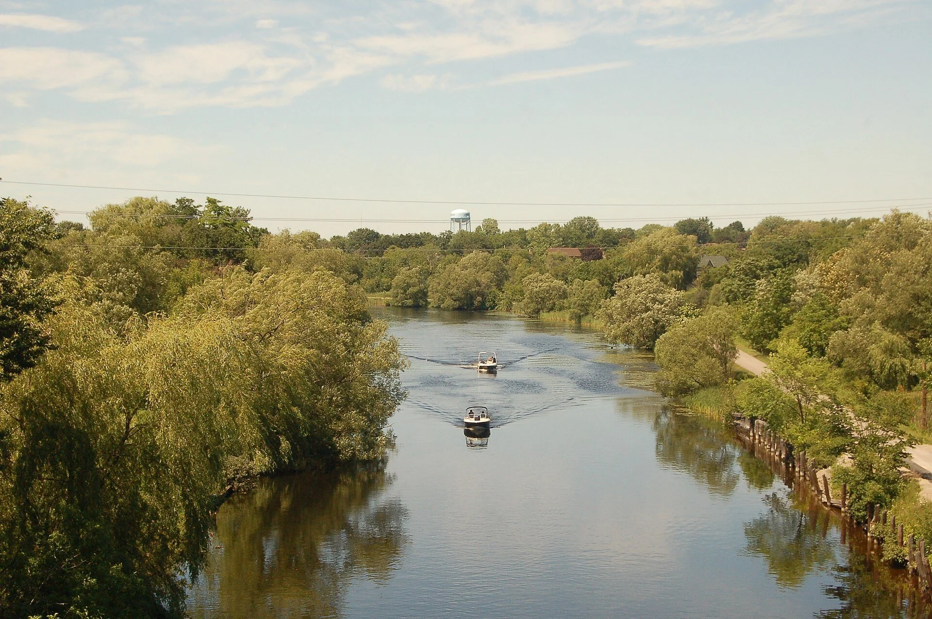 A small boat travelling up the tree-lined Scugog River in Lindsay, Ontario, in summer.
