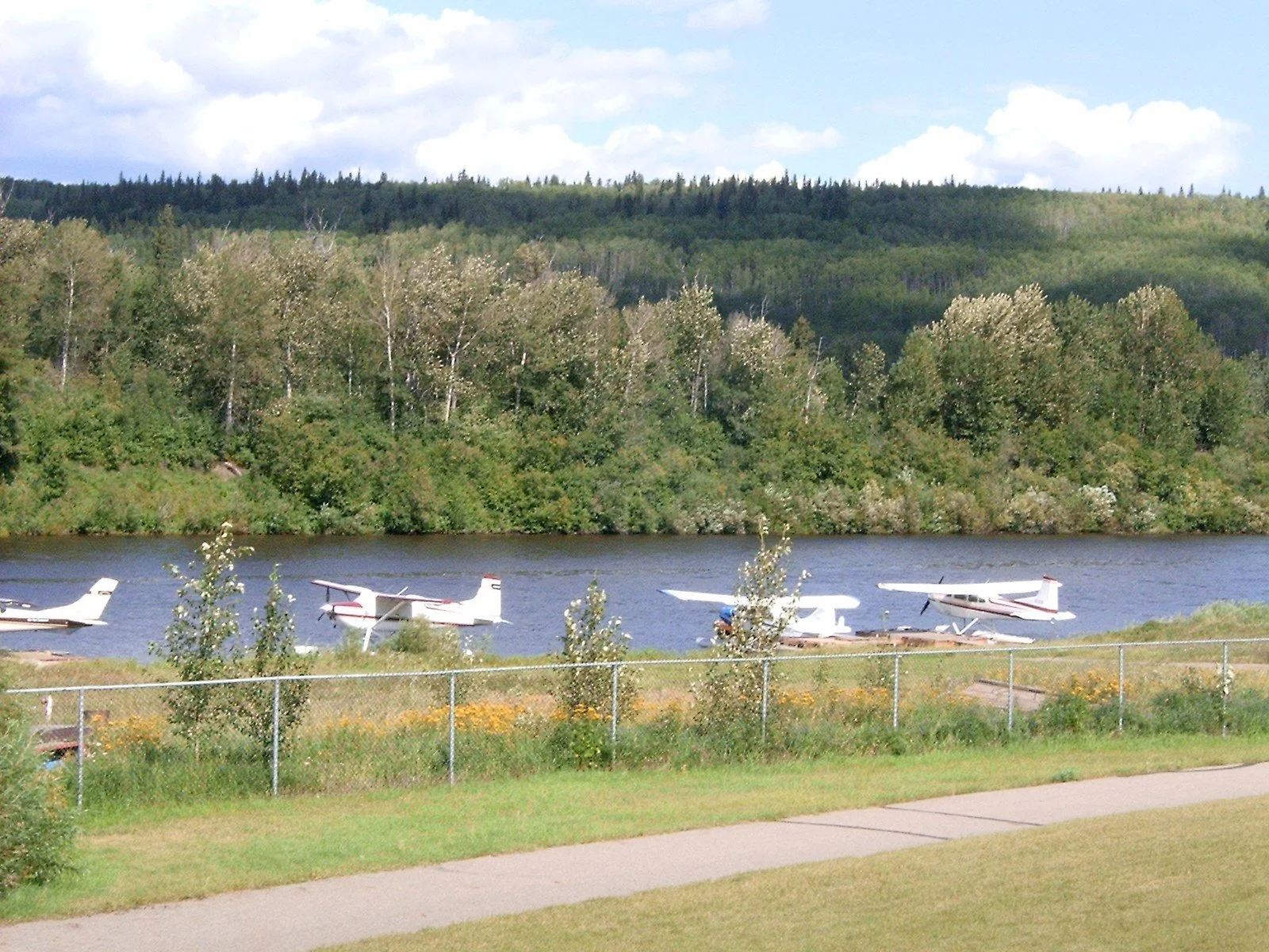 Float planes moored on the Snye channel in Fort McMurray, Alberta, with a paved path in front and wooded hills behind.