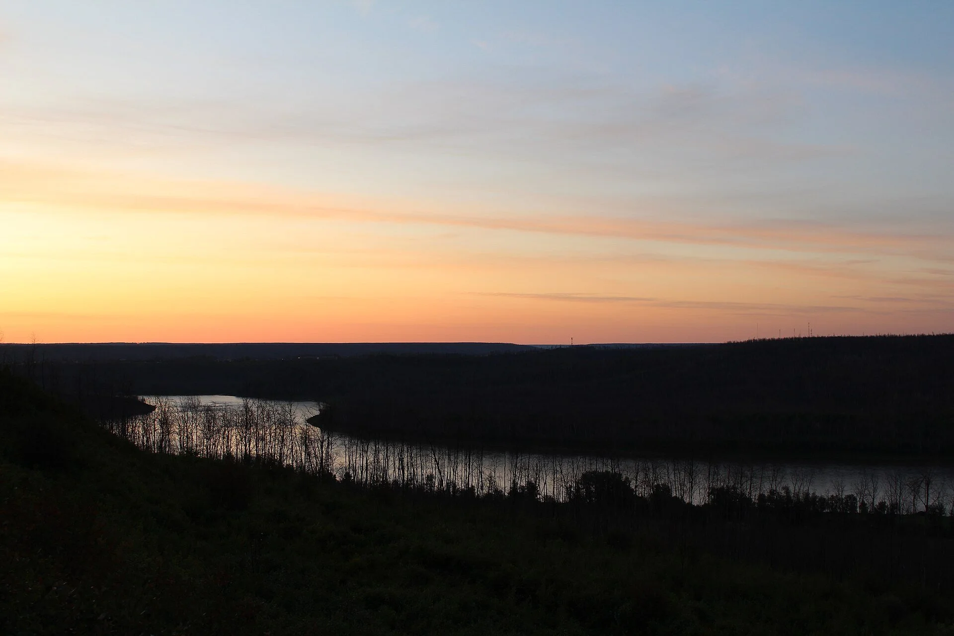 The Athabasca River winding through dark boreal forest under an orange sunrise sky in Fort McMurray, Alberta.