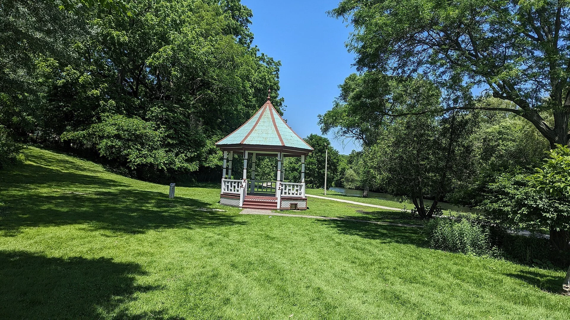 A white gazebo bandstand on a green lawn among tall trees in Lynnwood Park, Simcoe, Ontario.