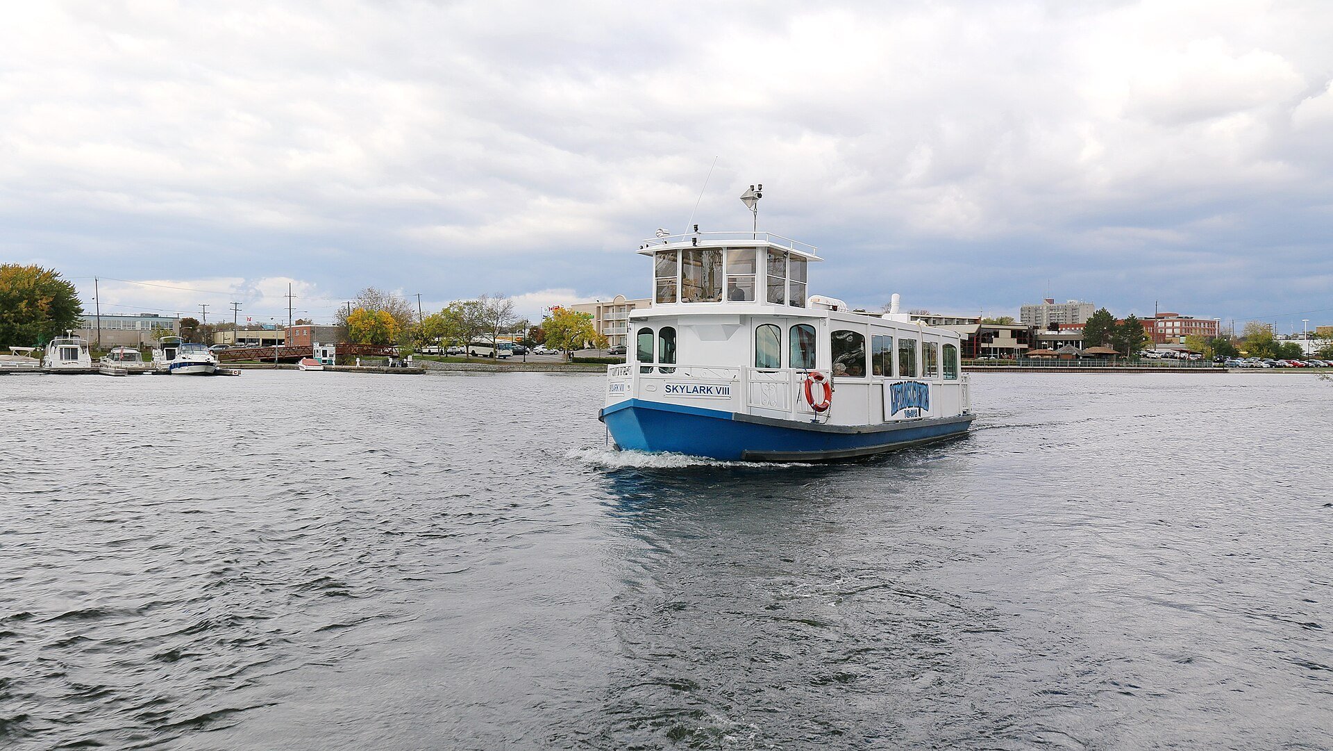 A small white and blue tour boat cruising across Little Lake with downtown Peterborough's shoreline behind it.
