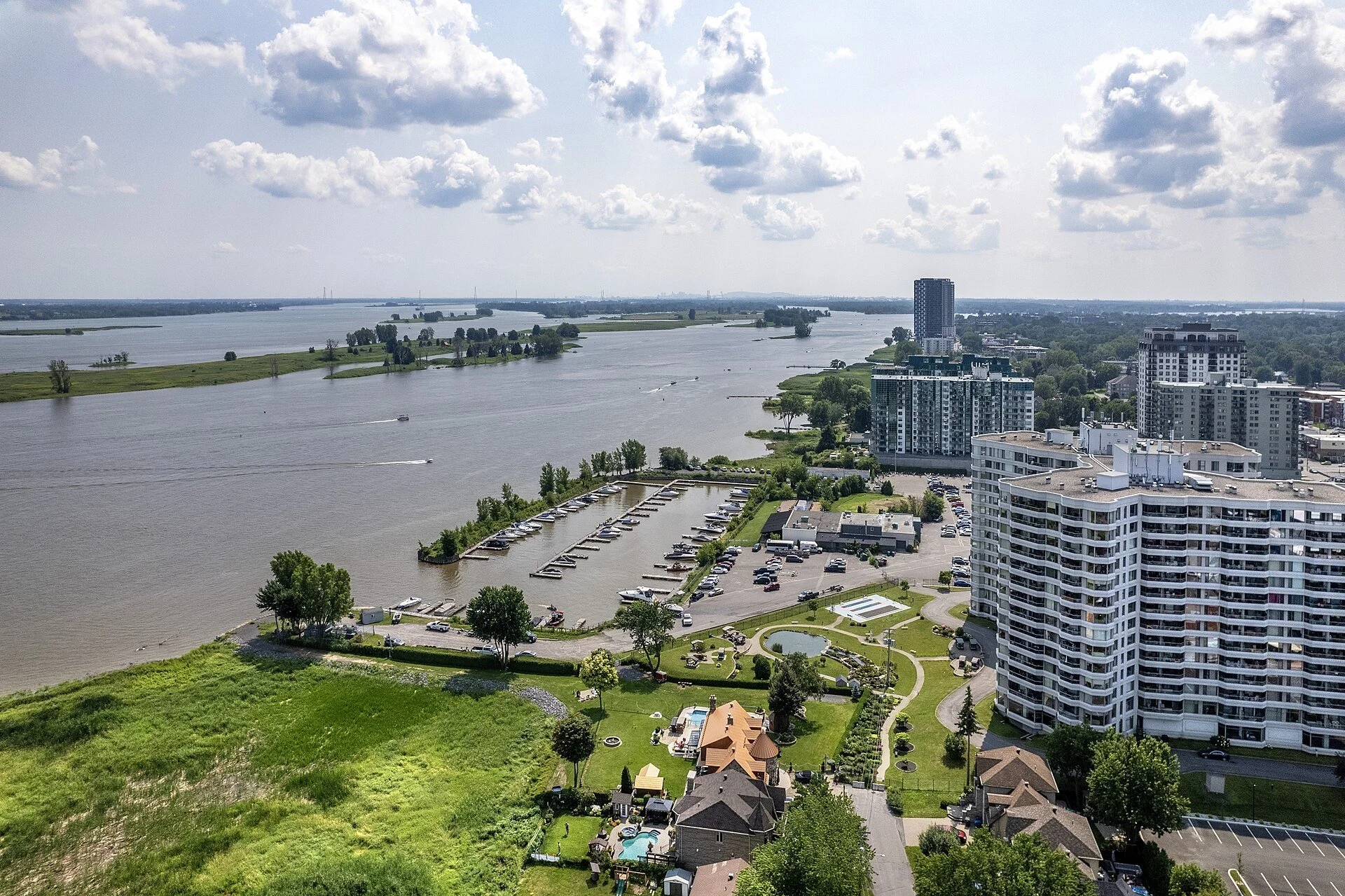 Aerial view of Repentigny's riverfront, with a marina, apartment towers and the broad St. Lawrence River under a cloudy sky.