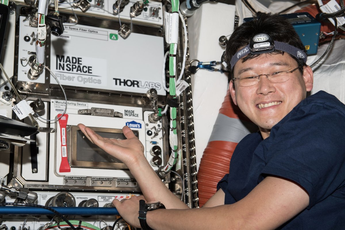 Japanese astronaut Norishige Kanai poses in front of Made In Space's Additive Manufacturing Facility (bottom) and Fiber Optics payload (top) which were installed next to each other during the company's recent fiber optics mission.