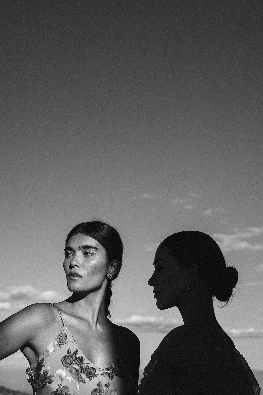 Two women, one with a floral dress and the other with a bun hairstyle, standing outdoors against a partly cloudy sky in black and white.