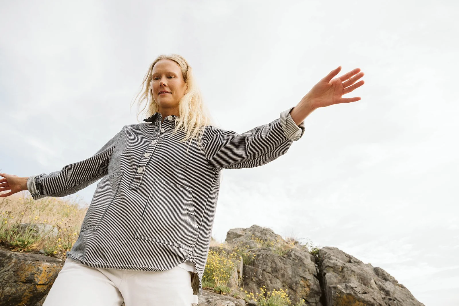 A woman with blonde hair in a striped button-up shirt and white pants stands outdoors with arms extended to the sides, standing on a rocky landscape with small flowers and a cloudy sky in the background.
