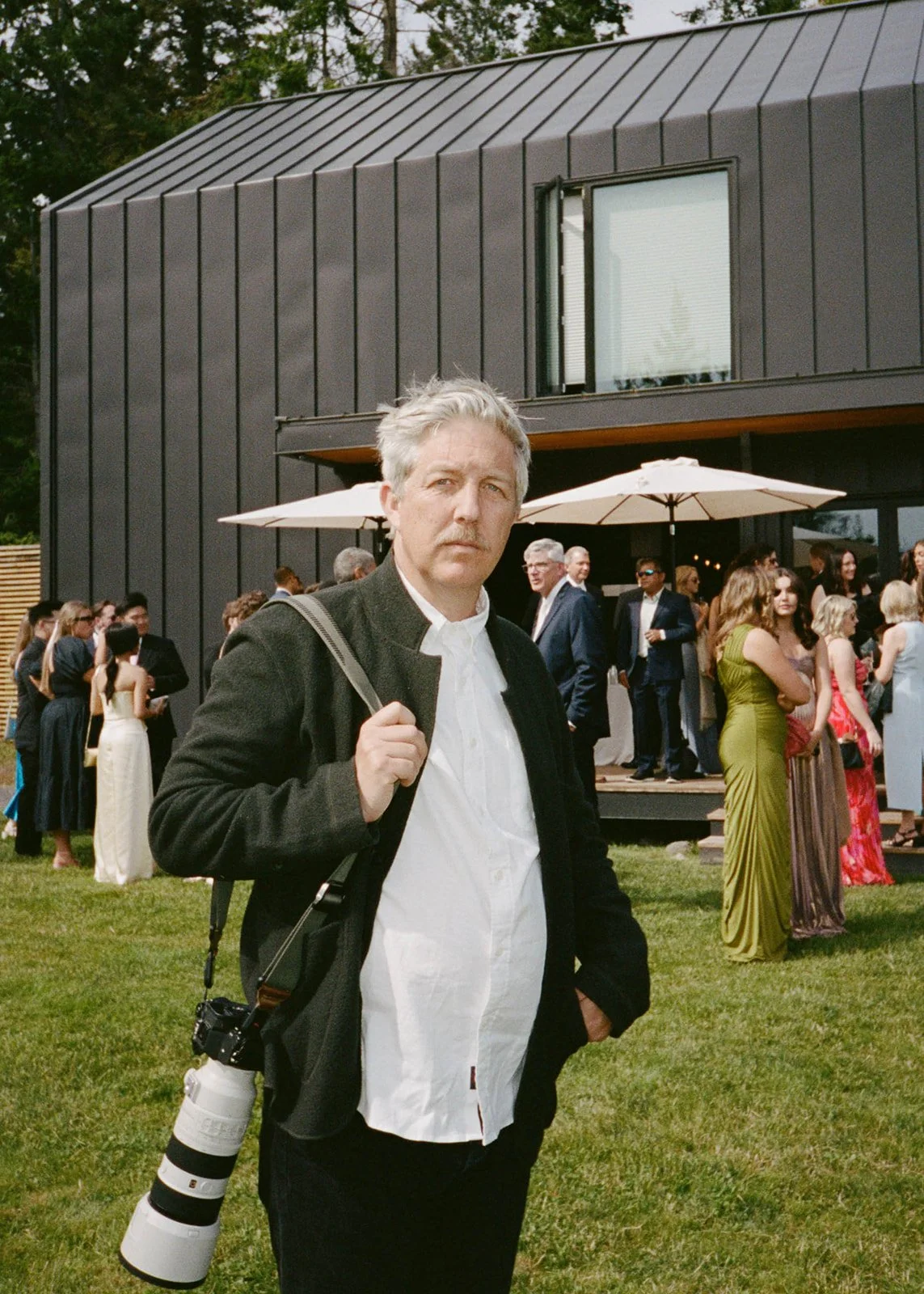 A man standing outdoors at a social gathering or party, holding a camera with a large telephoto lens, with a modern dark building and a crowd of people in elegant attire in the background.