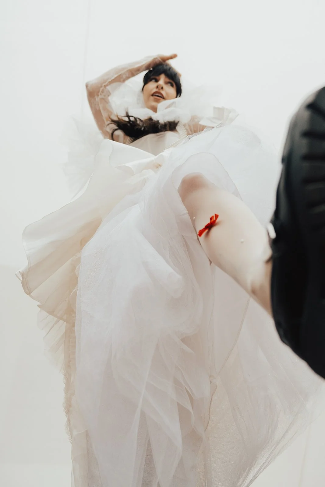 A woman dressed in a white gown with ruffles and lace details, lying on her back and looking up, with a medical tourniquet on her arm, and a photographer taking the picture from below against a plain background.