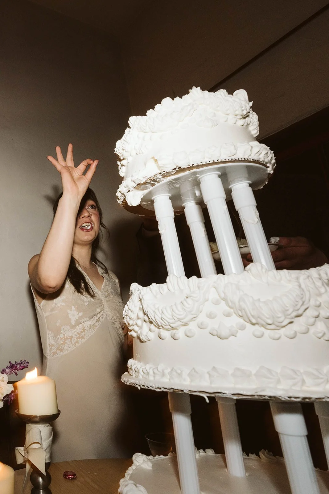 A woman with dark hair and a beige lace dress celebrating next to a large, tiered white cake with columns and decorative icing.