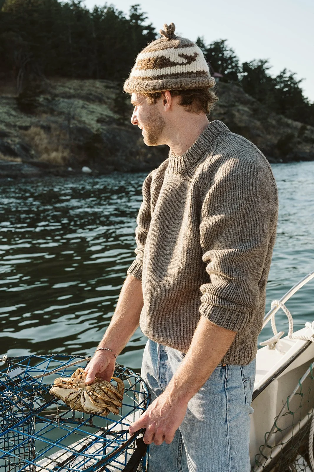 Man with a beige sweater, jeans, and a knitted hat holding a crab on a fishing trap on a boat by the water, with trees and rocky land in the background.