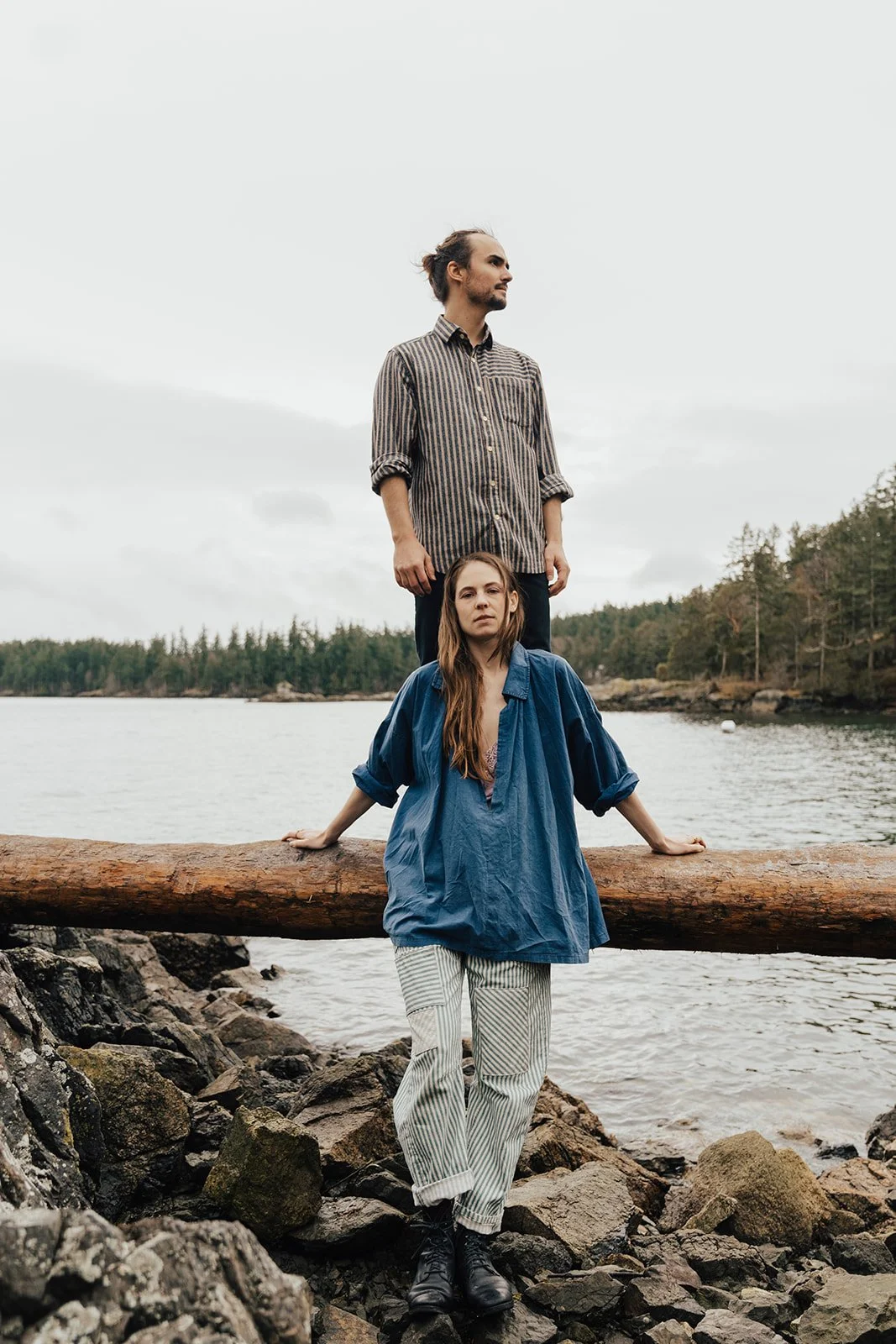 A man and a woman near a body of water with trees in the background. The woman is sitting on rocks with her hands on a log behind her, and the man is standing behind her on the log. Overcast sky.