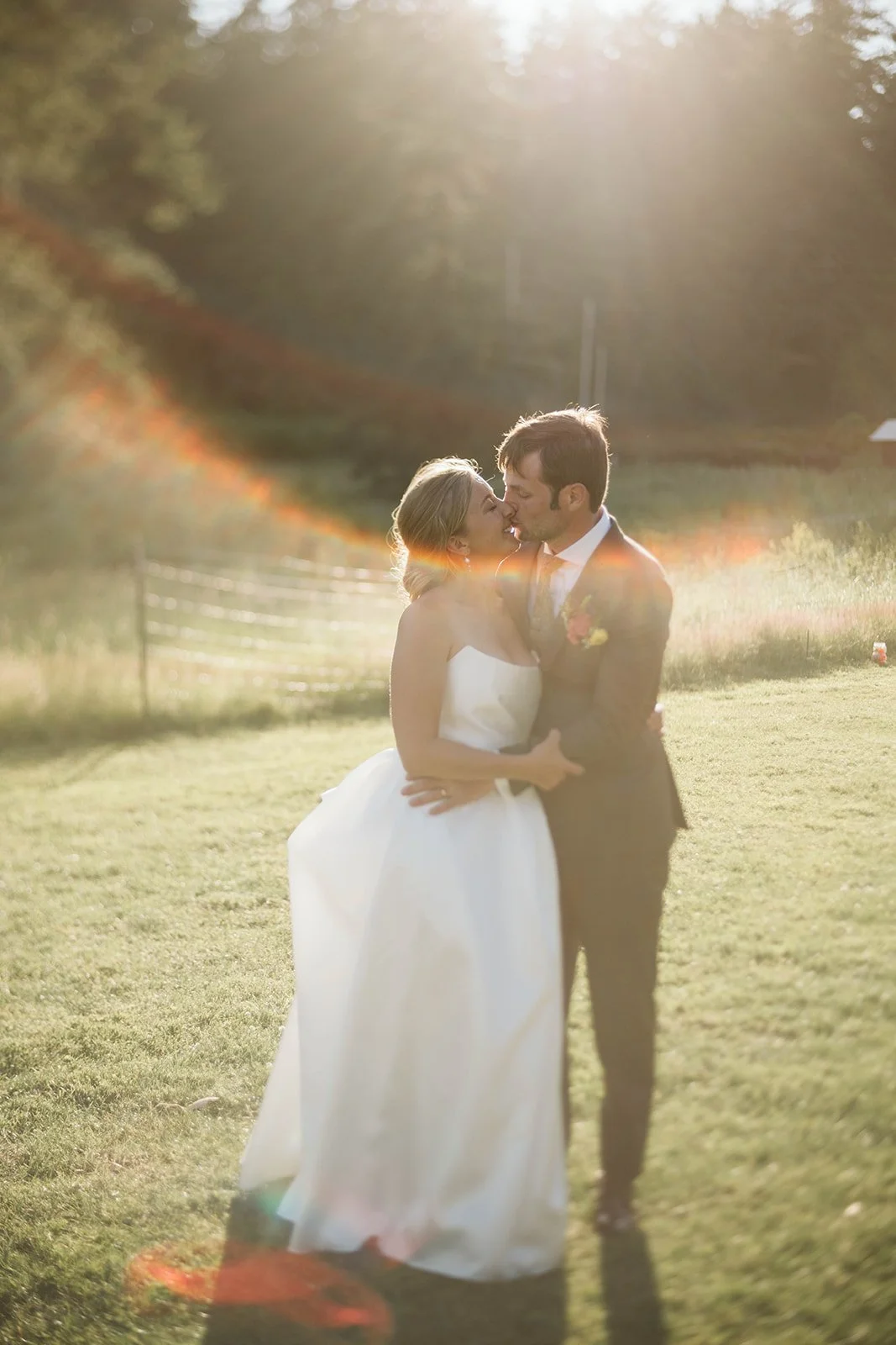 A bride and groom sharing a kiss outdoors during sunset, with sunlight creating a lens flare.