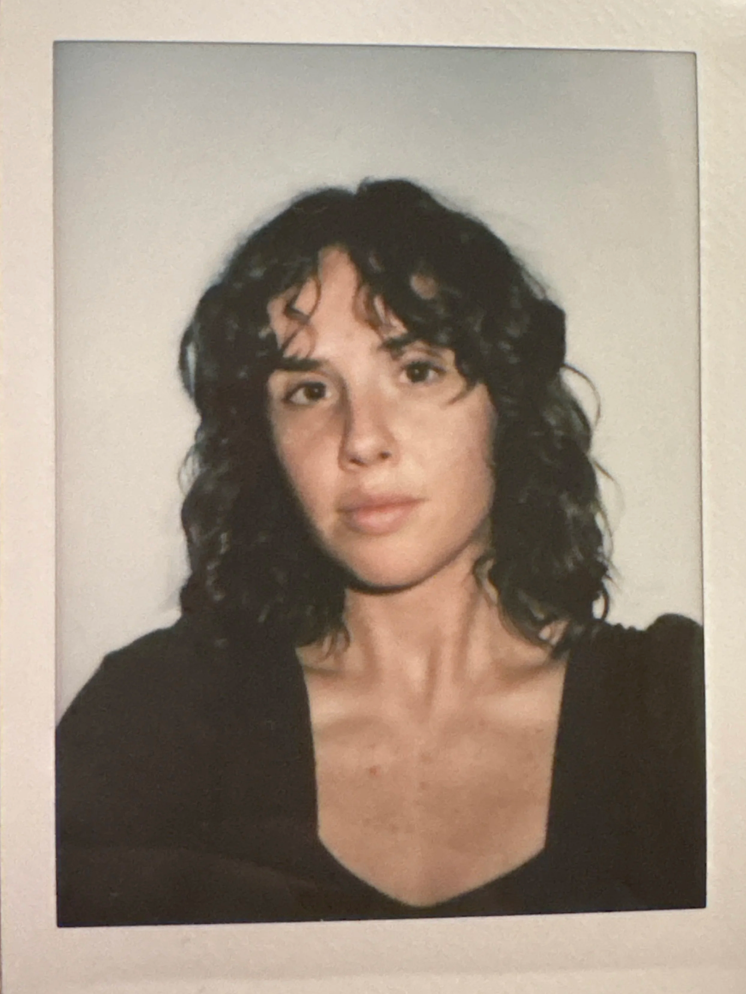 A woman with shoulder-length curly black hair, wearing a black top, looking directly at the camera with a neutral expression, standing against a plain light background.
