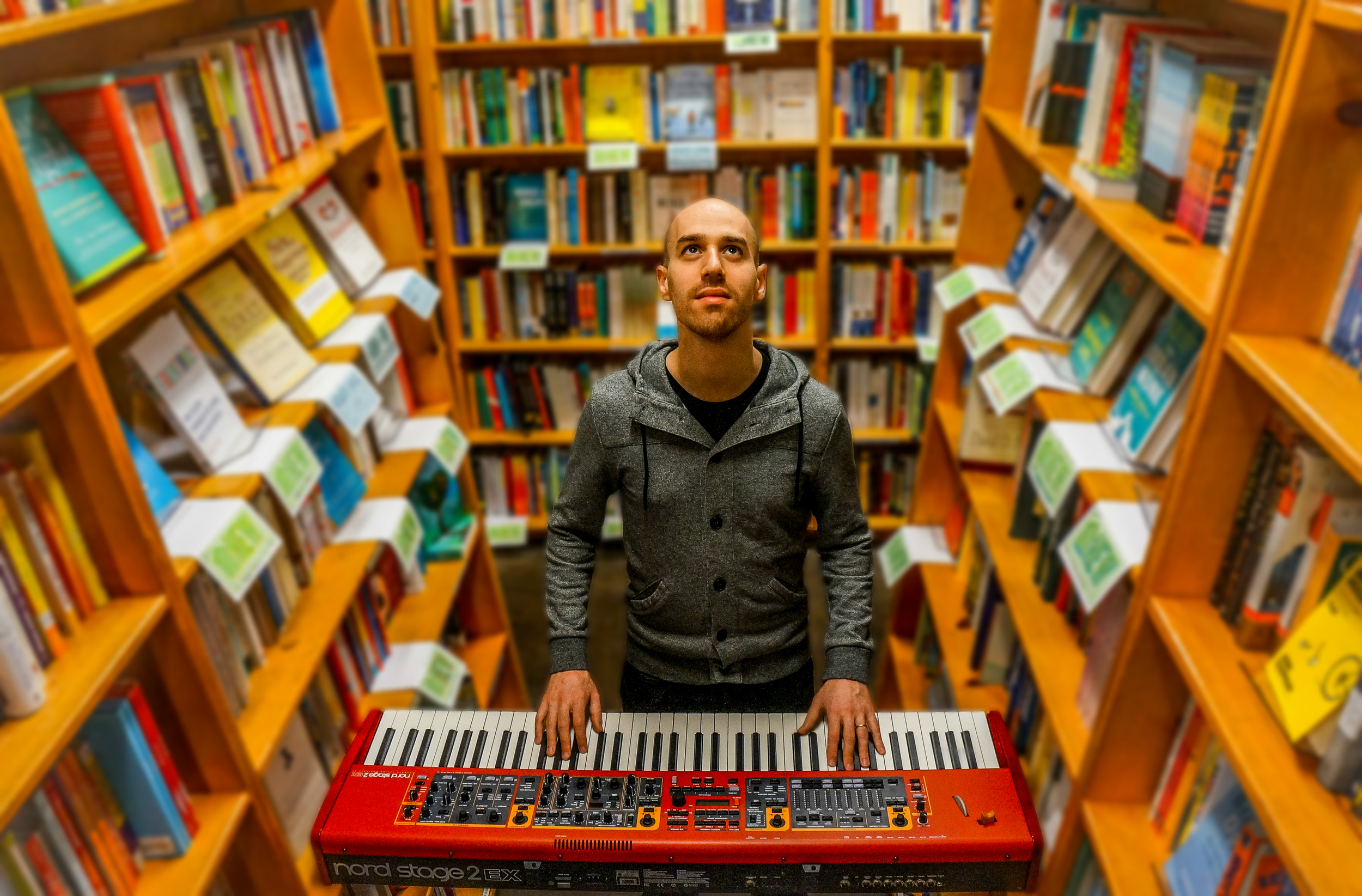 Man in grey hoodie playing red keyboard between shelves of books in a bookstore.