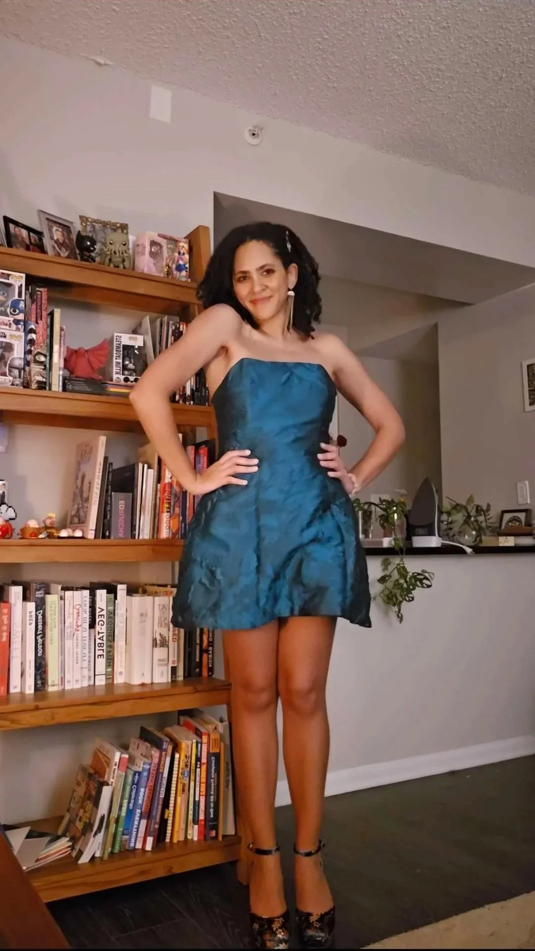A black woman wearing a teal dress poses in front a bookcase..