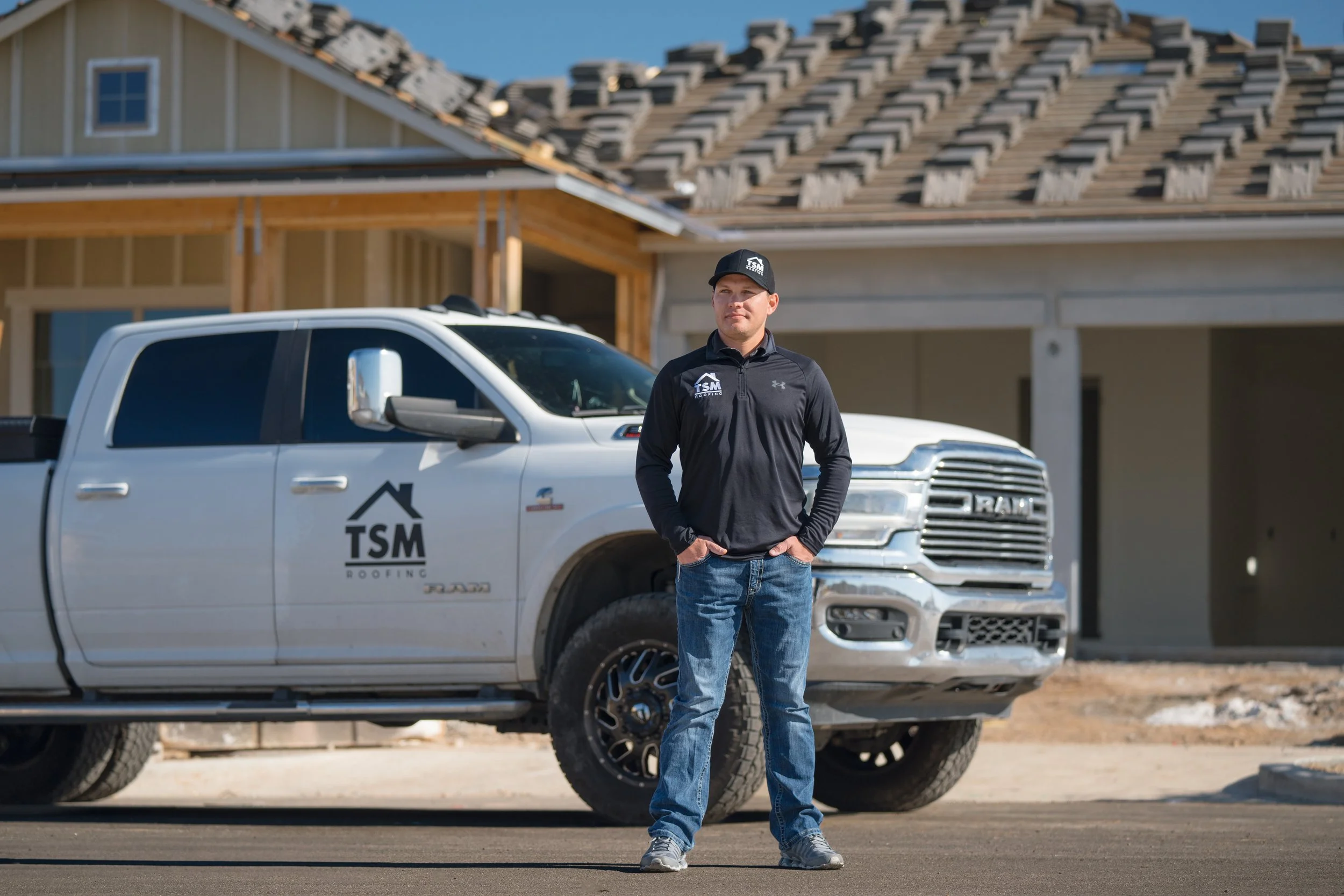 A man standing in front of a construction site with a white RAM truck with TSM Roofing logo parked behind him.