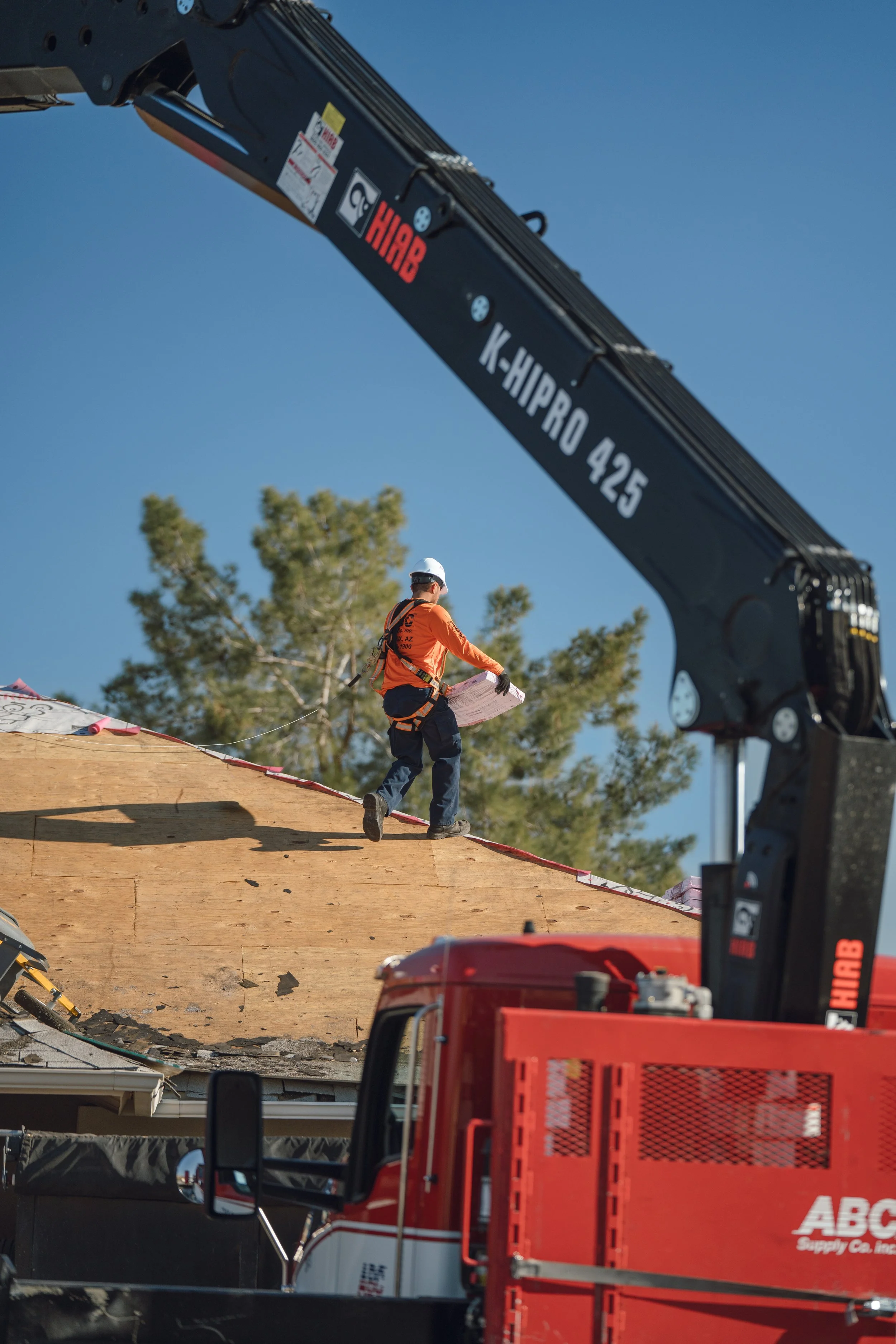 Construction worker in safety gear walking on a sloped wooden roof surrounded by construction equipment, with a large crane and trees in the background under a clear blue sky.