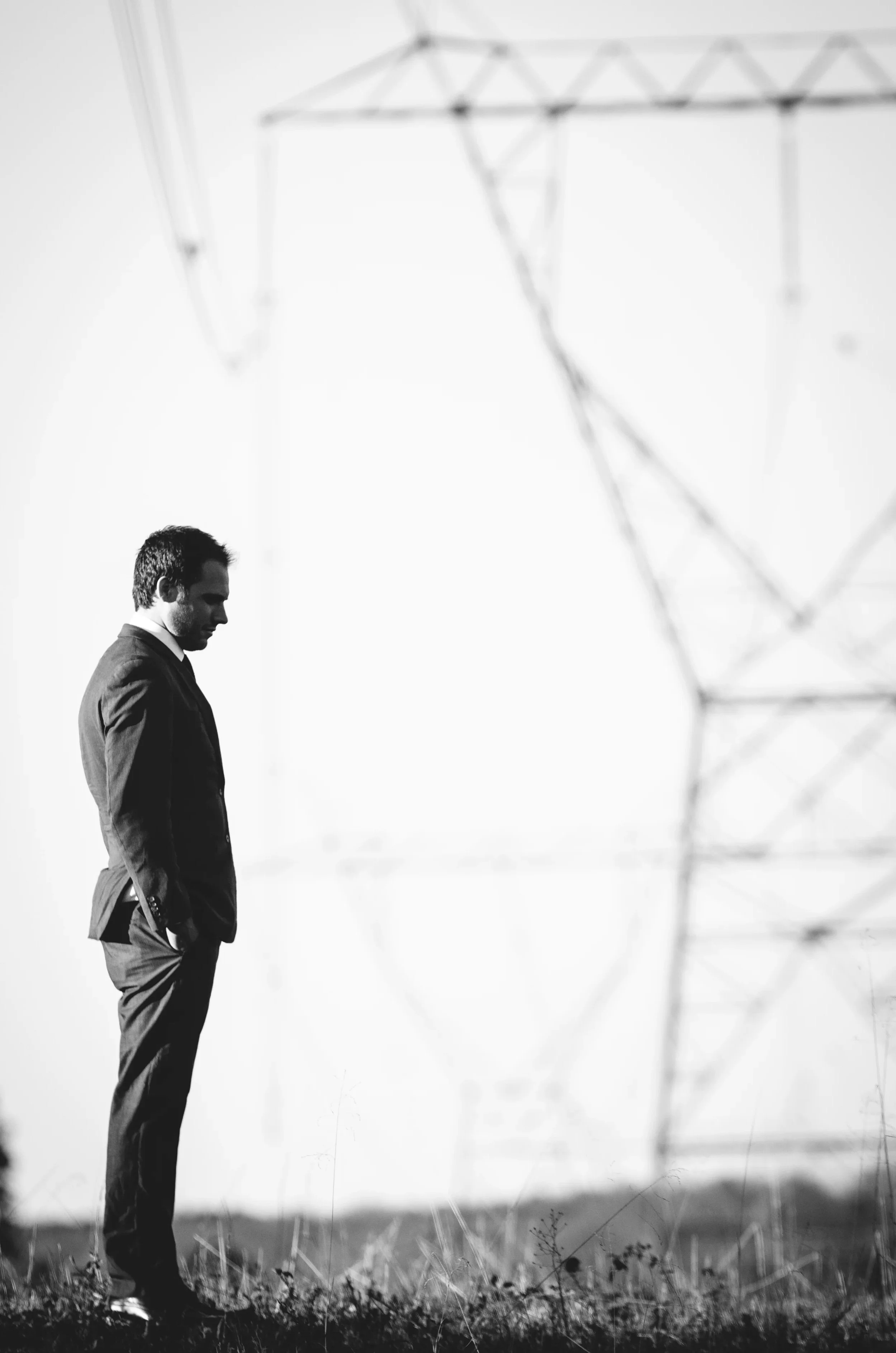 Black and white photo of a man in a suit standing in a field with a large electricity pylon in the background.