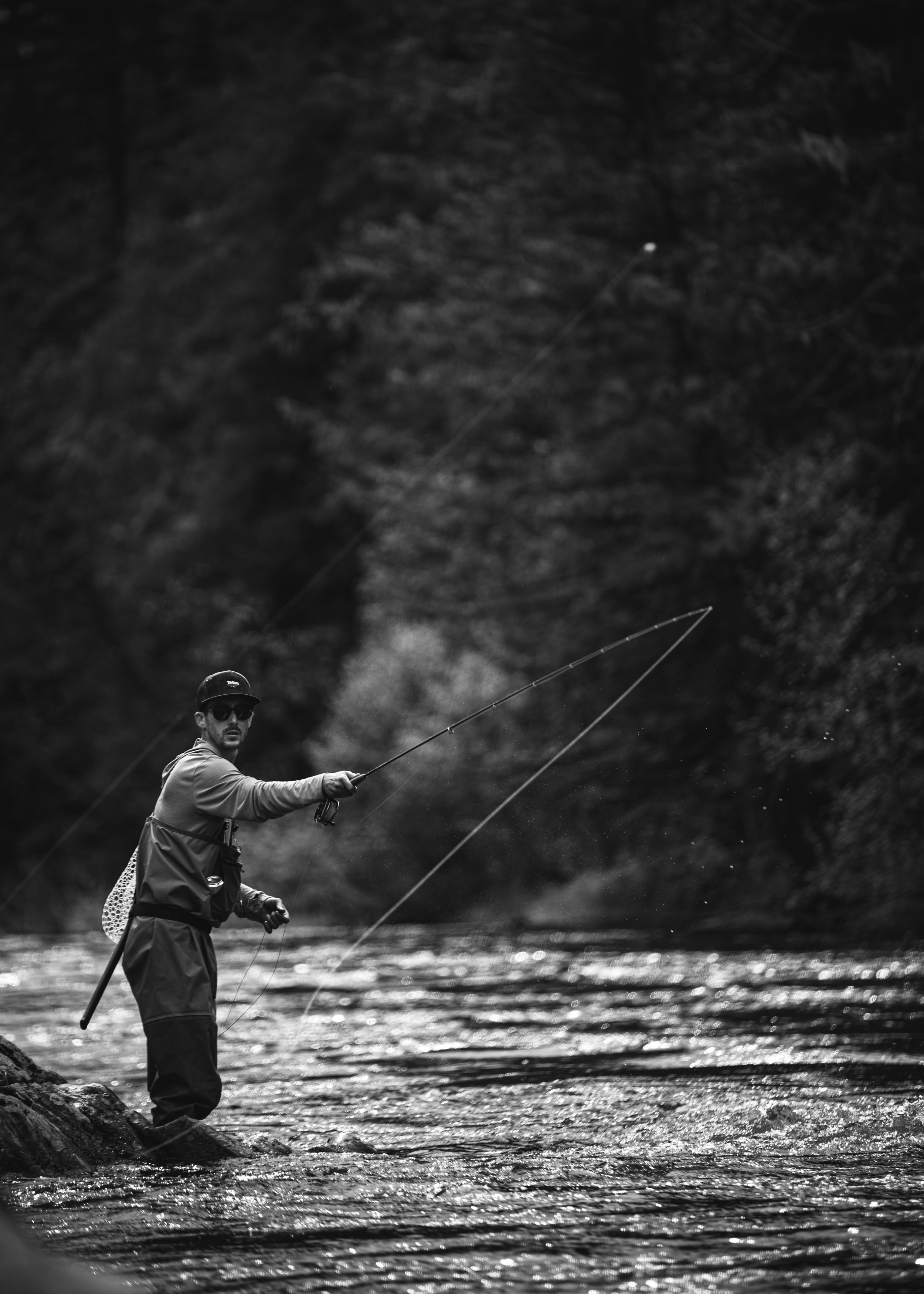 A man fishing in a river, standing on rocks, with trees in the background.