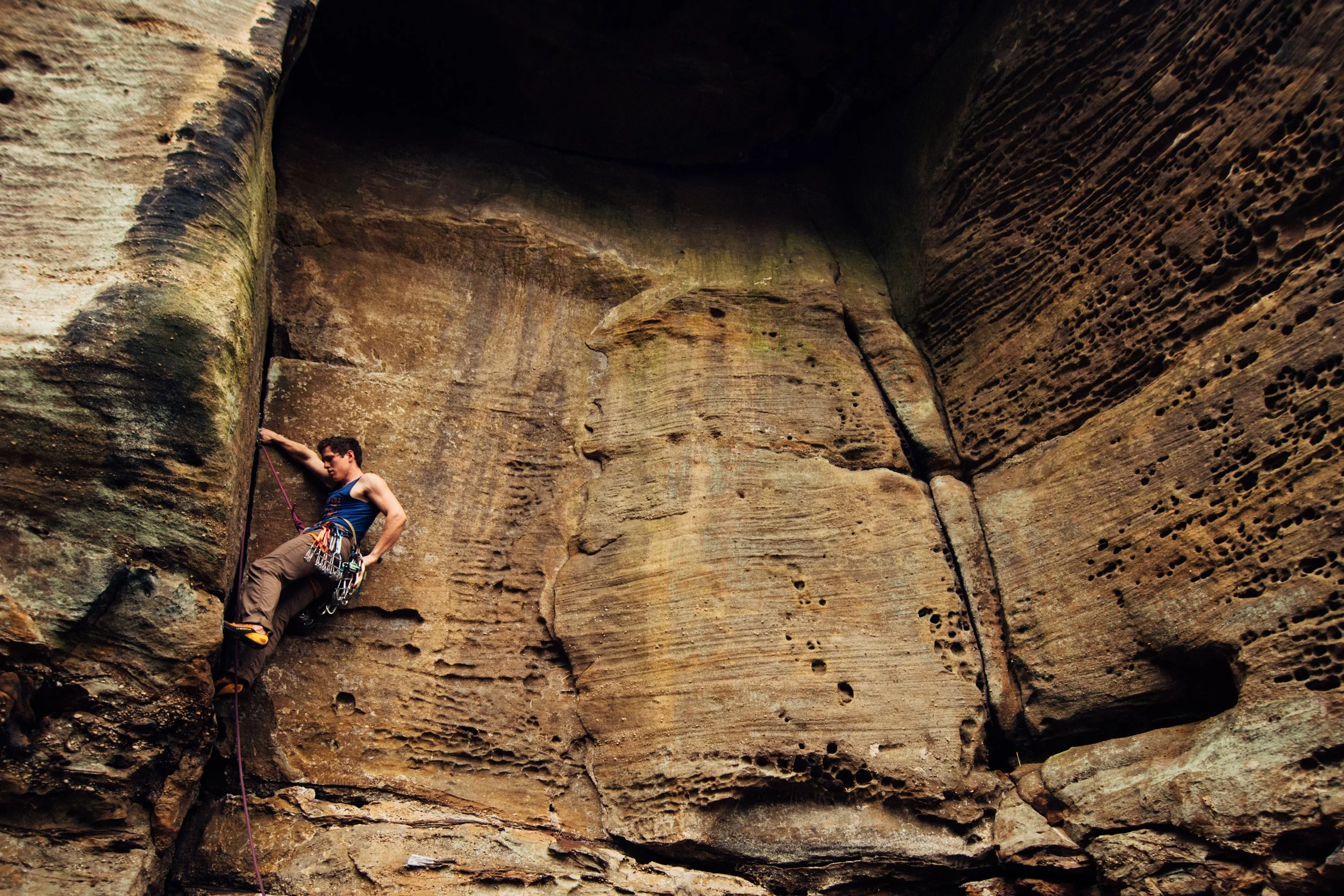 A man rock climbing on a large vertical rock face with rough textured sandstone surface and small holes, inside a natural canyon or cliff face.
