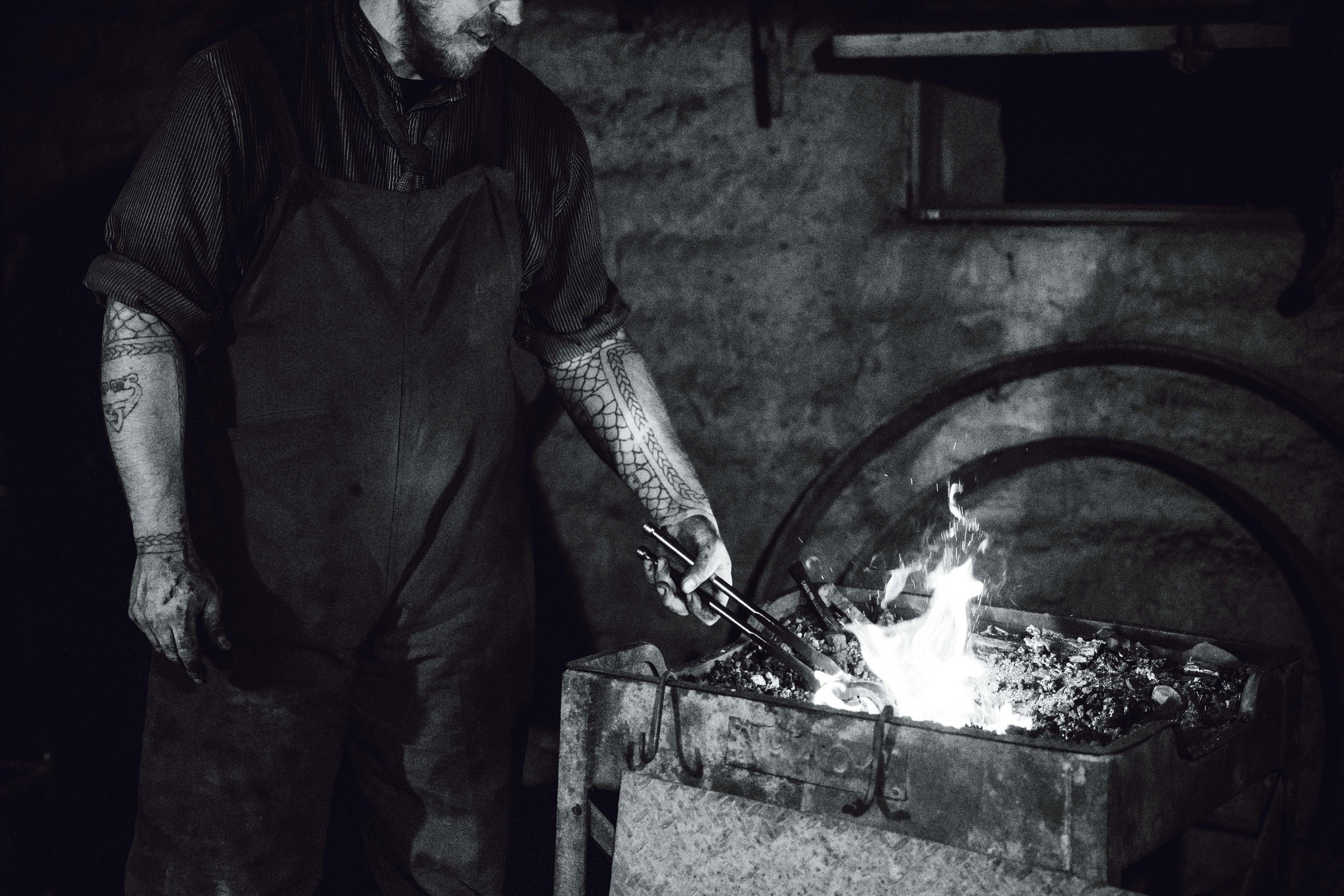 A person with tattoos wearing a dark apron black shirt, working at a forge with a bright flame in a workshop.