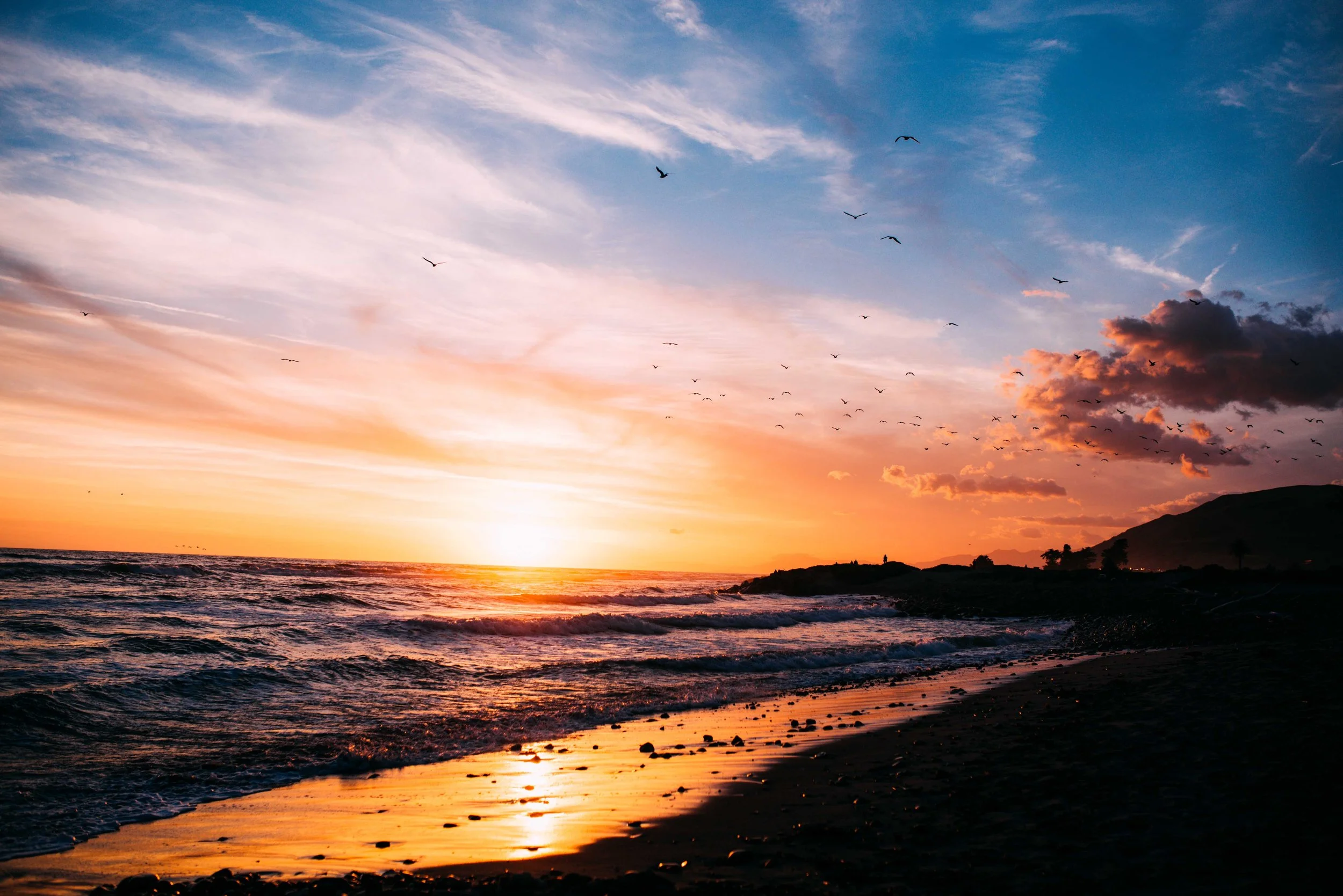 Sunset at the beach with the ocean, silhouetted hills, flying birds, and colorful skies.