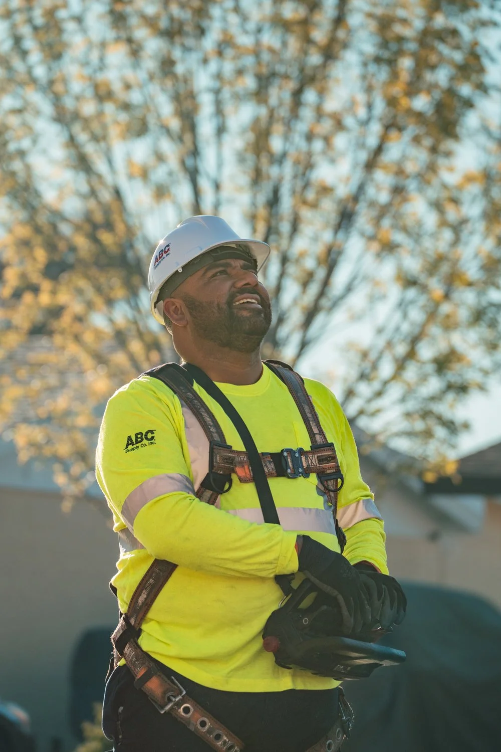 A construction worker wearing a helmet and high-visibility yellow shirt, smiling and looking up, standing outdoors with trees and a building in the background.