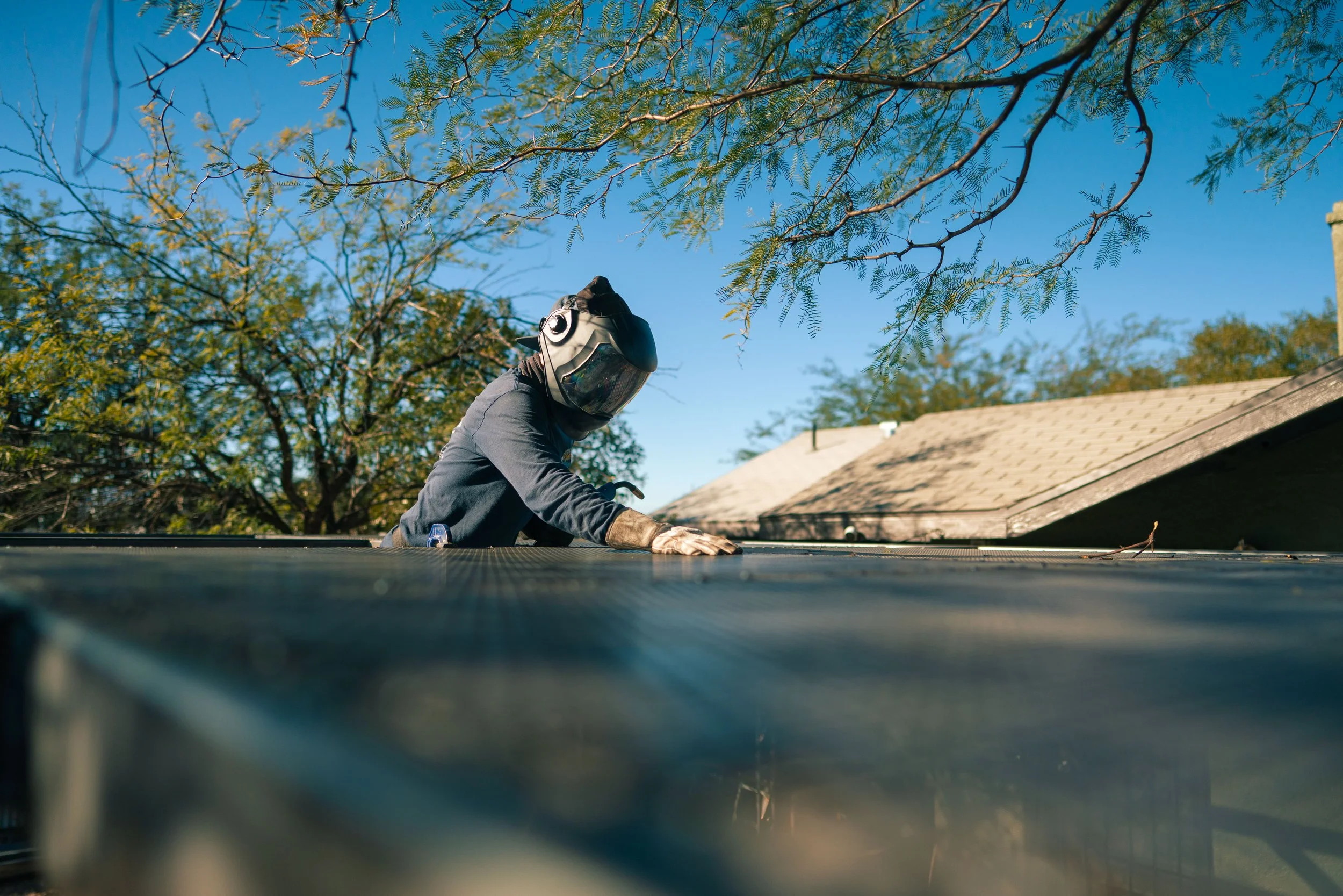 A worker wearing a helmet, gloves, and protective clothing is on a roof, working under a blue sky and trees with changing leaves.