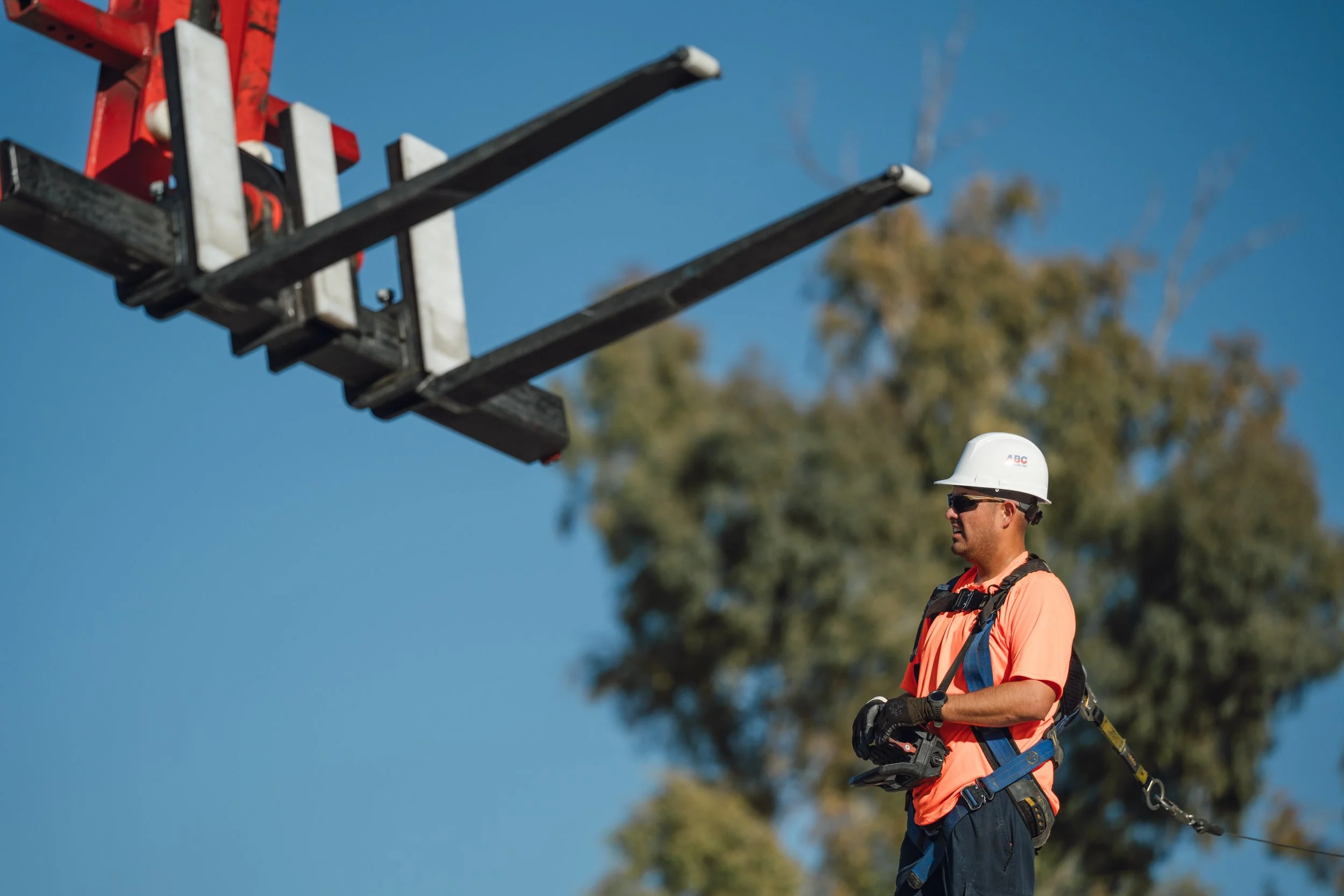A construction worker wearing a white helmet, sunglasses, an orange shirt, and a safety harness standing outdoors under a blue sky with trees in the background.